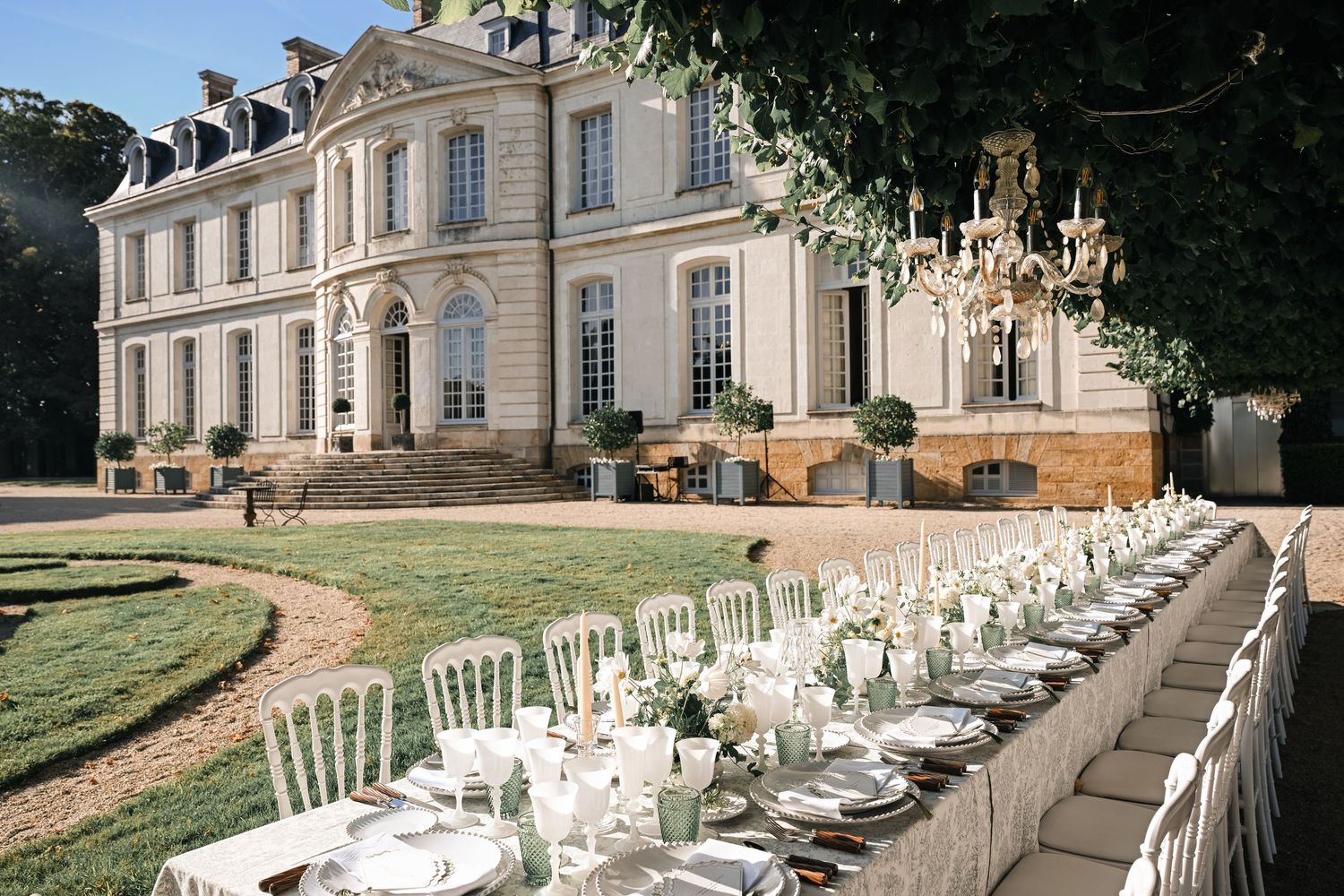 Luxury French chateau wedding reception with long outdoor dining table and chandelier.