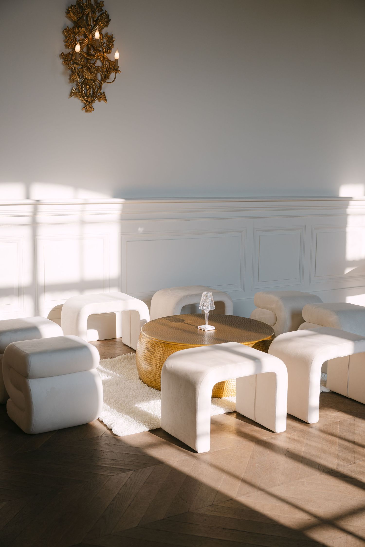 Bright interior with modern white curved chairs around a gold drum coffee table beneath an ornate gilded sconce.