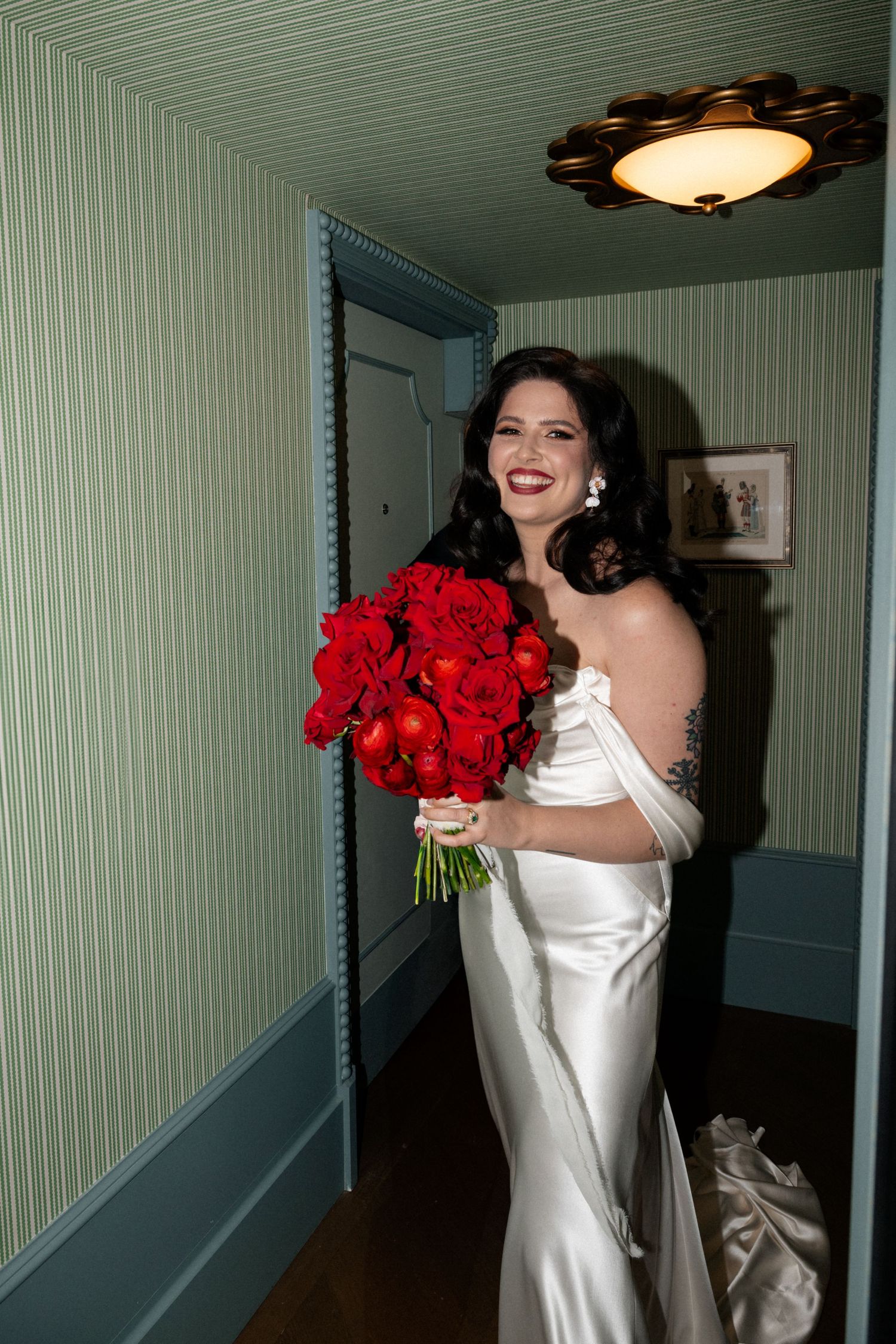 Bride in white satin gown holding vibrant red rose bouquet in hallway.