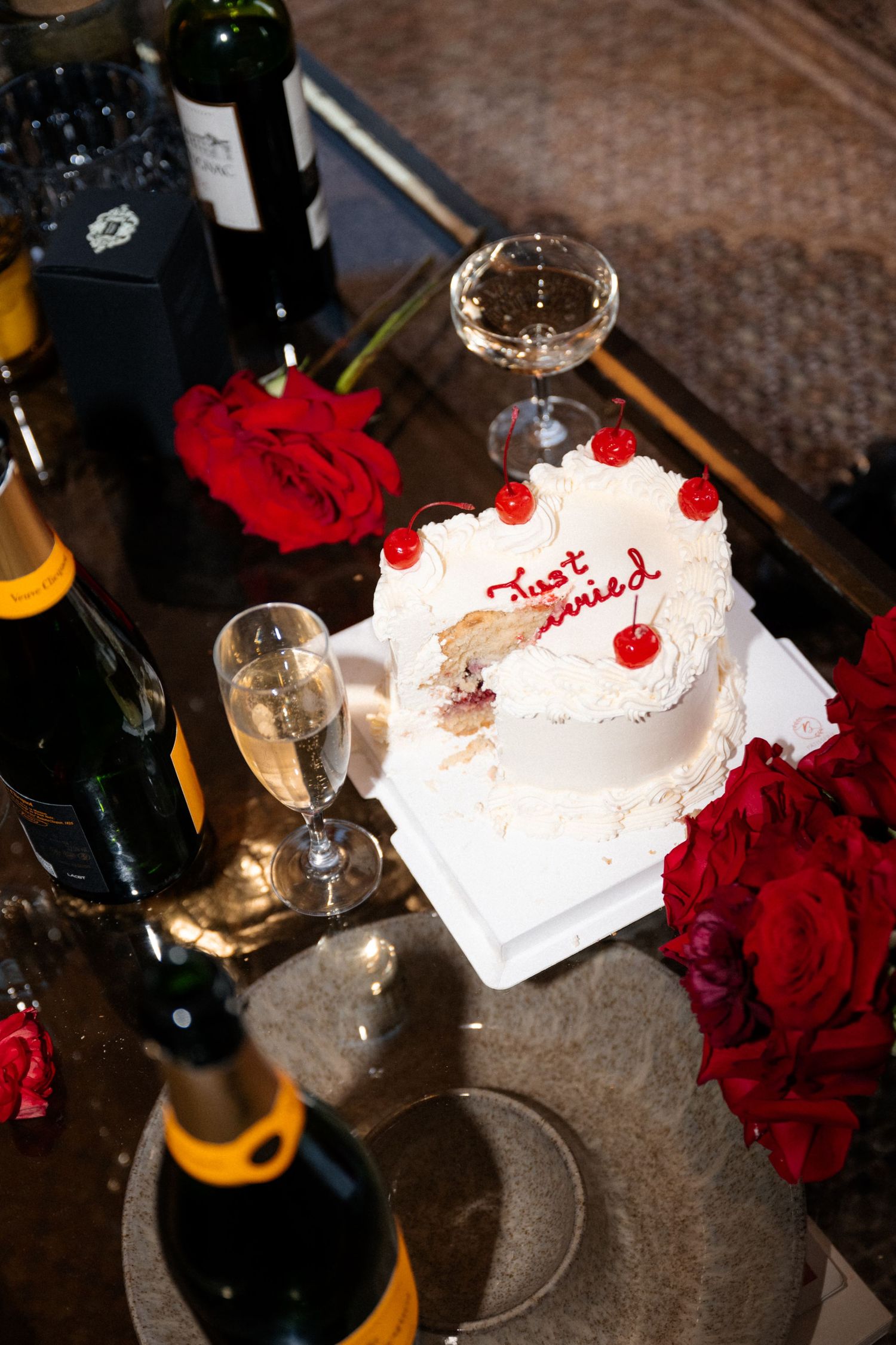 Just Married heart-shaped white wedding cake with cherries beside Veuve Clicquot champagne and red roses.