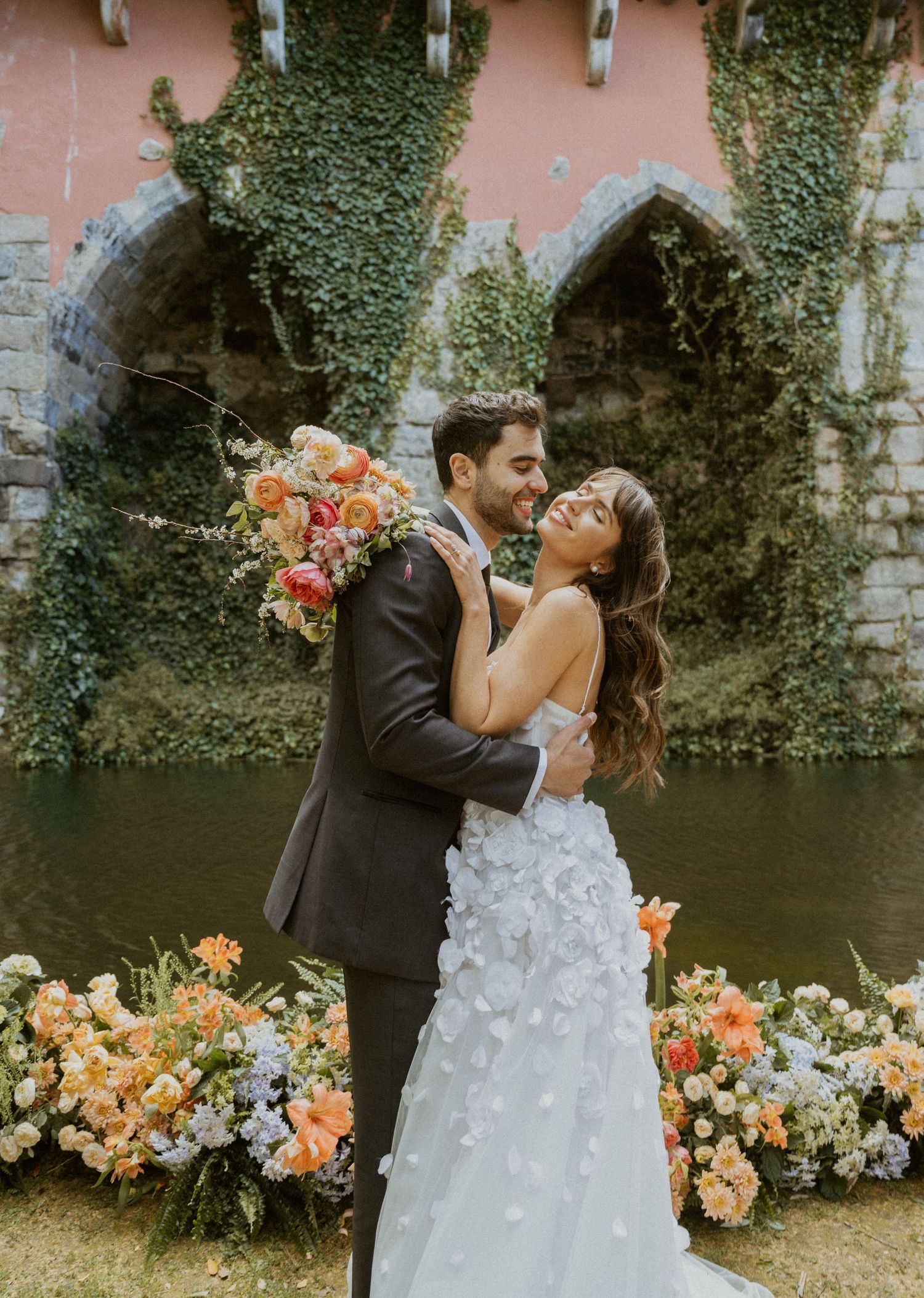 A couple embraces in wedding attire surrounded by colorful floral arrangements before an ivy-covered stone bridge.