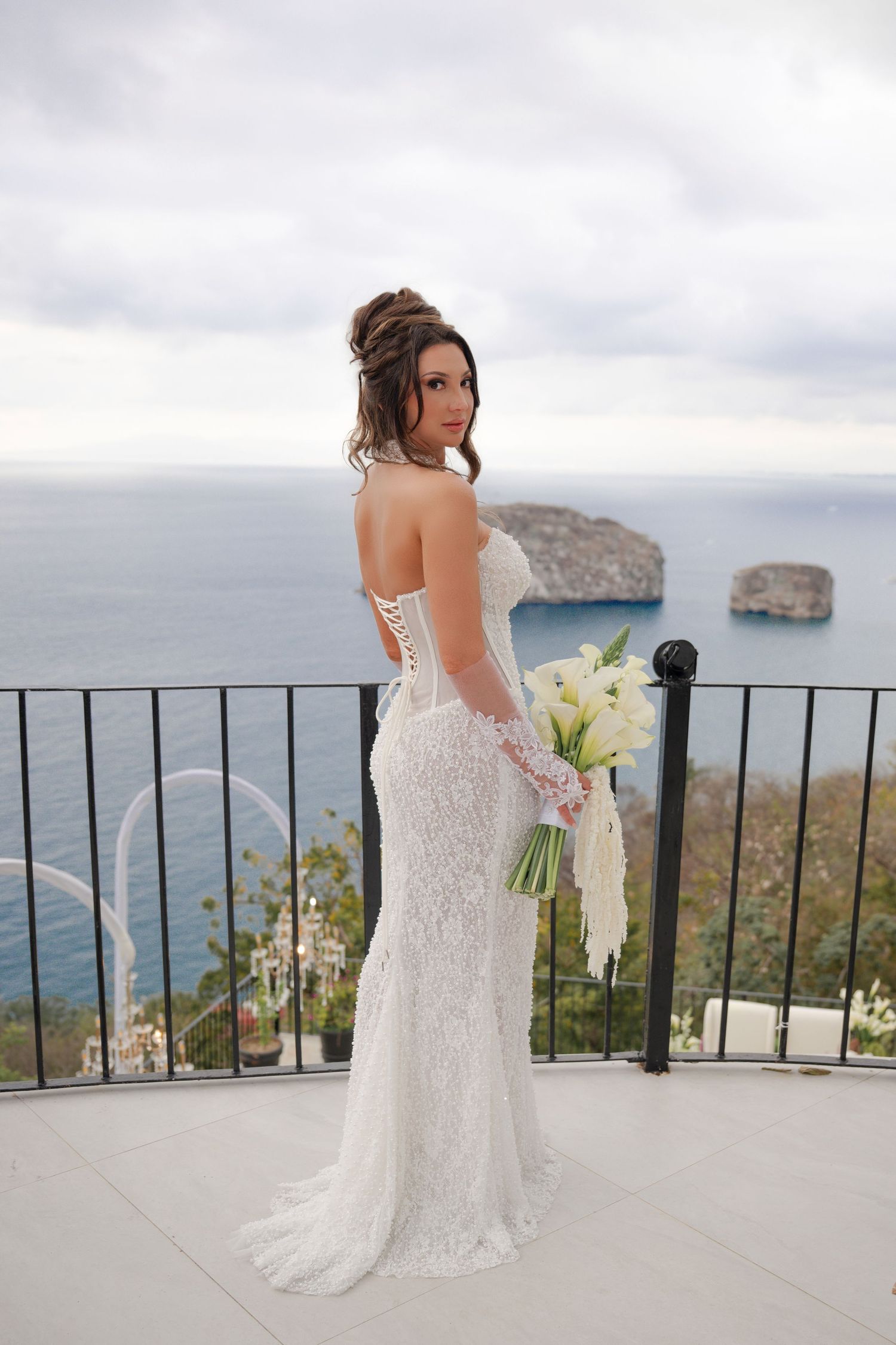 Elegant bride in white lace corset gown holding bouquet on seaside balcony with ocean view.