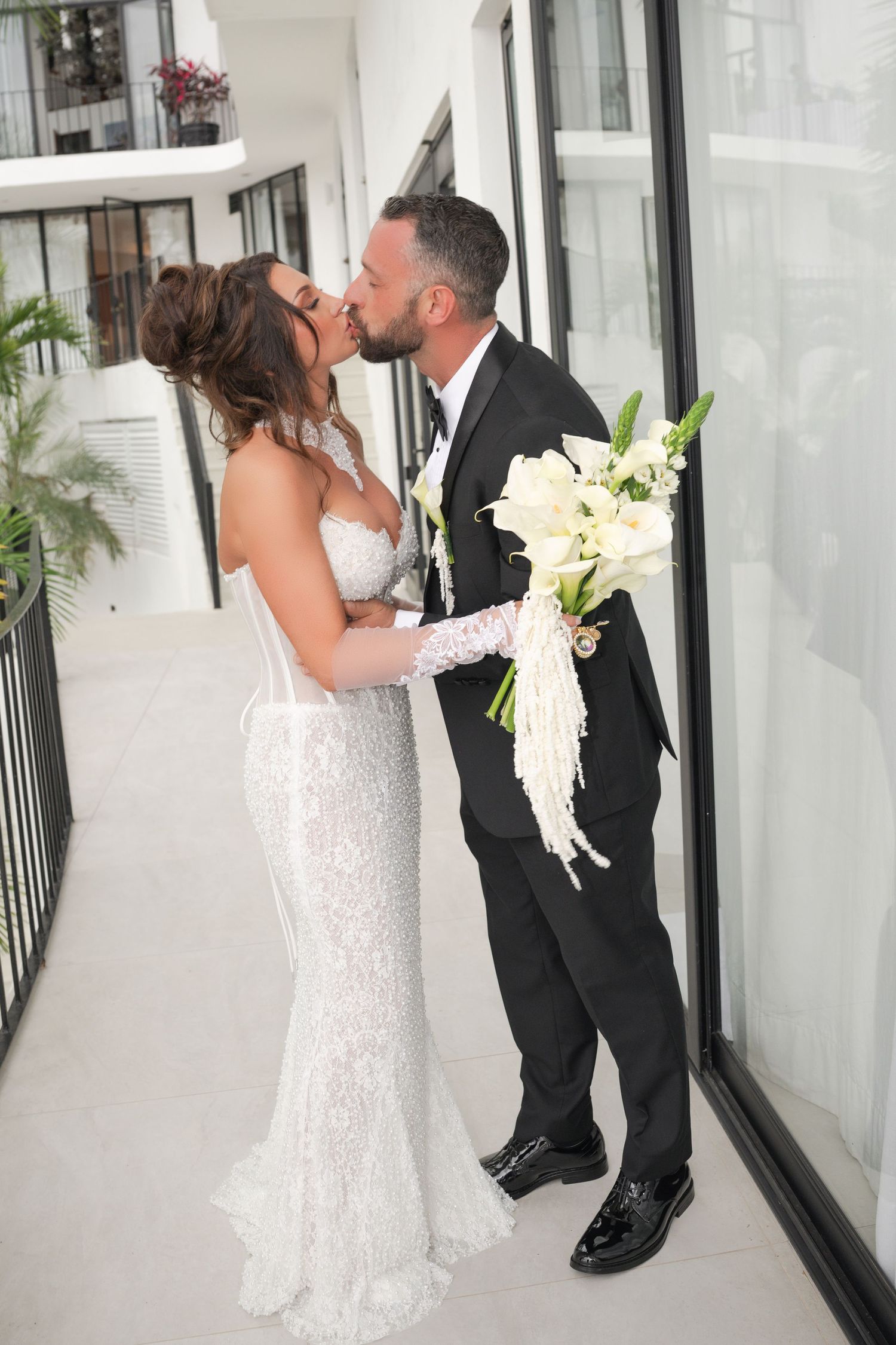 Bride and groom share a kiss on a balcony, she holds white calla lily bouquet wearing lace gown, he wears black tuxedo.
