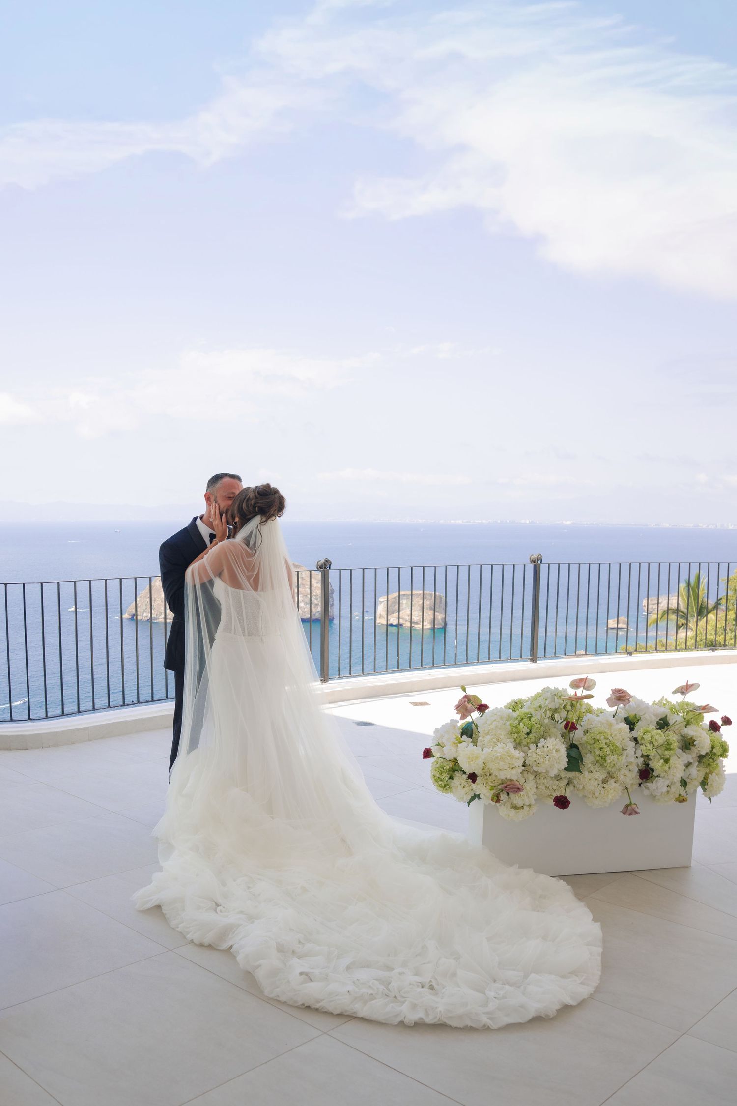 Bride and groom share a kiss on a scenic terrace overlooking the blue sea, with elegant white floral arrangements nearby.