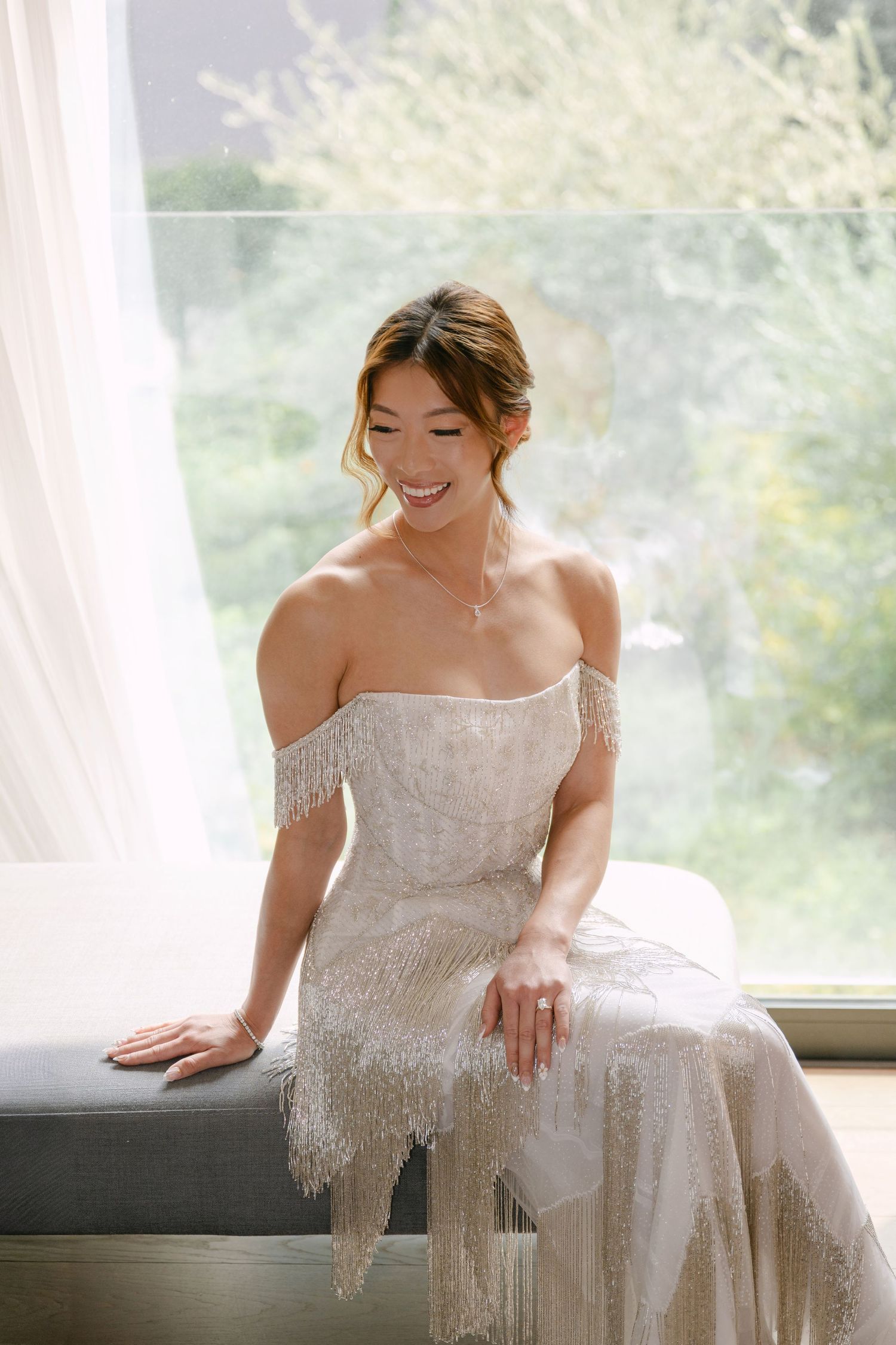 Smiling bride in off-shoulder beaded fringe wedding gown seated by a sunlit window with green trees in background.