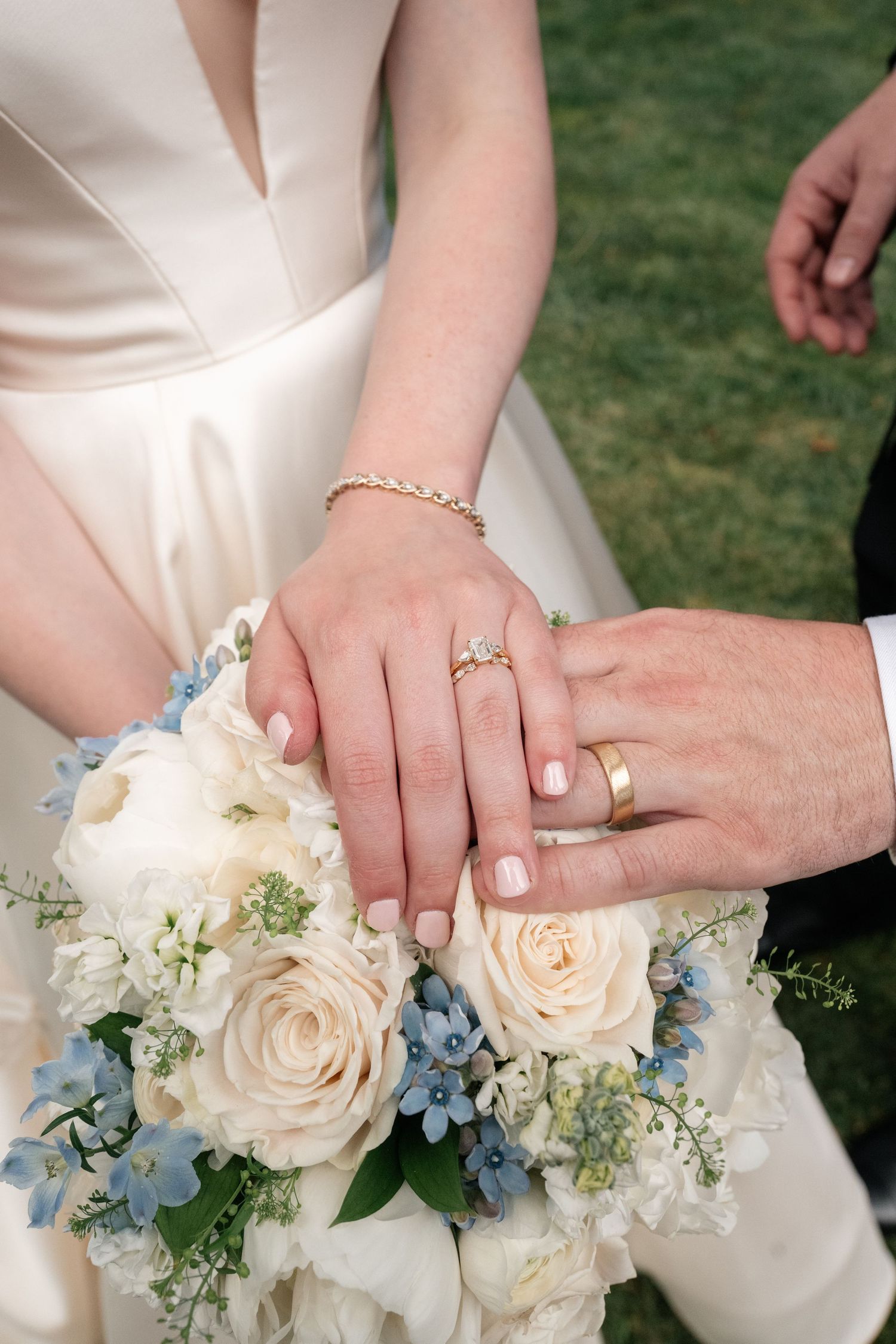 Bride and groom display wedding rings resting hands on white and blue bridal bouquet of roses.
