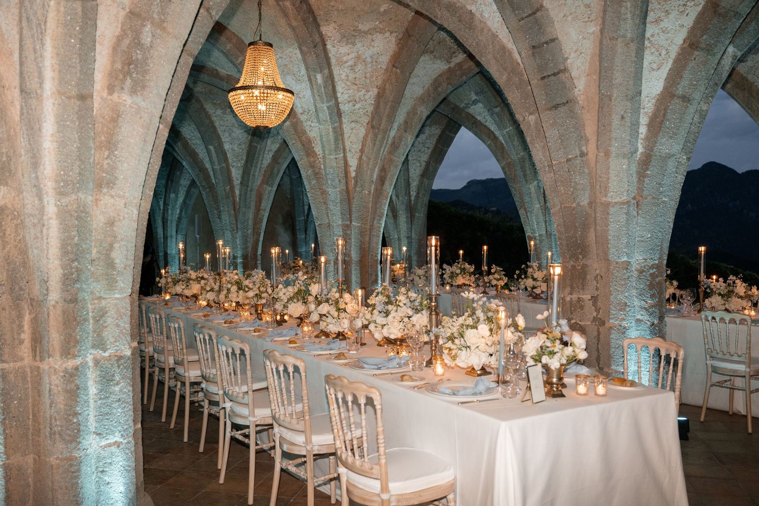 Elegant wedding reception in a medieval stone arched hall with long tables, candelabras, and floral centerpieces.