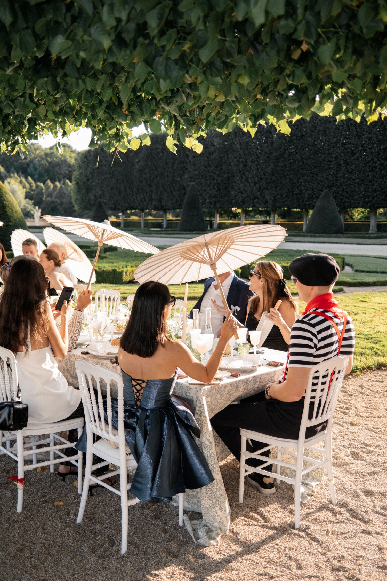 Guests dining al fresco under trees with parasols at an elegant outdoor garden party event.