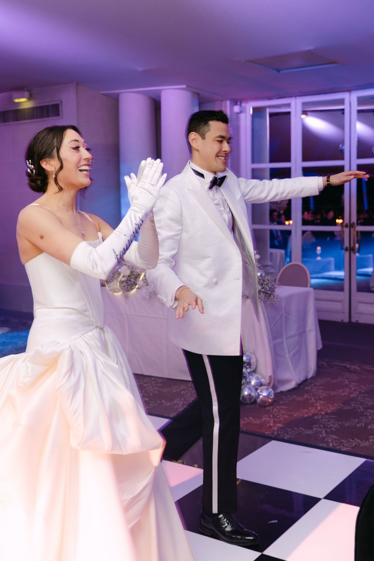 Smiling bride and groom in white tuxedo perform choreographed first dance on checkered floor.
