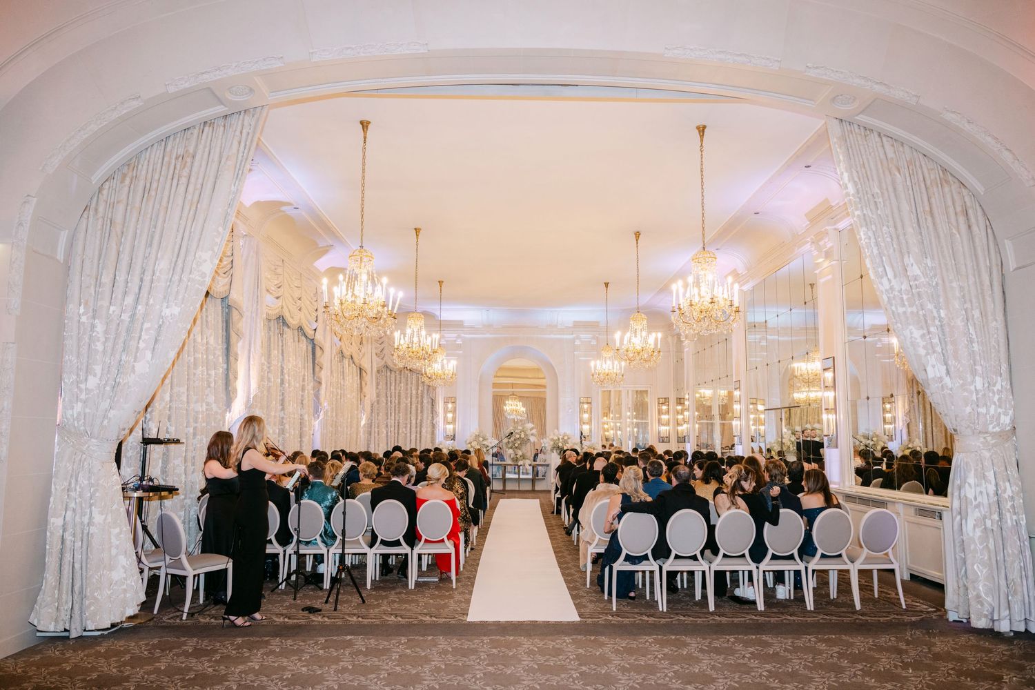 Elegant wedding ceremony in grand ballroom with crystal chandeliers, white aisle, and seated guests.