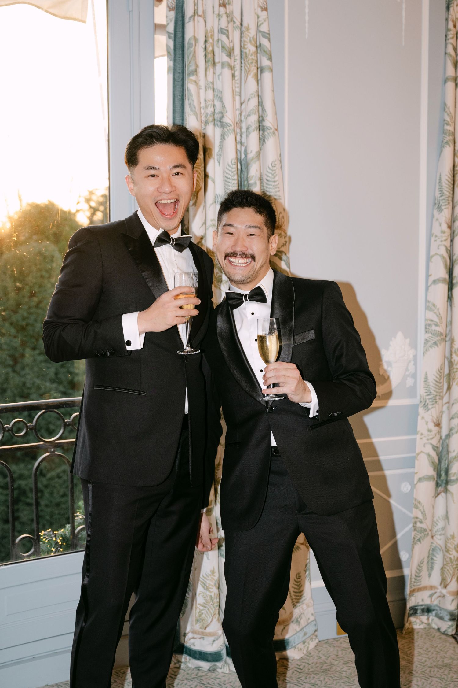 Two groomsmen in black tuxedos smiling and raising champagne glasses near sunlit window.