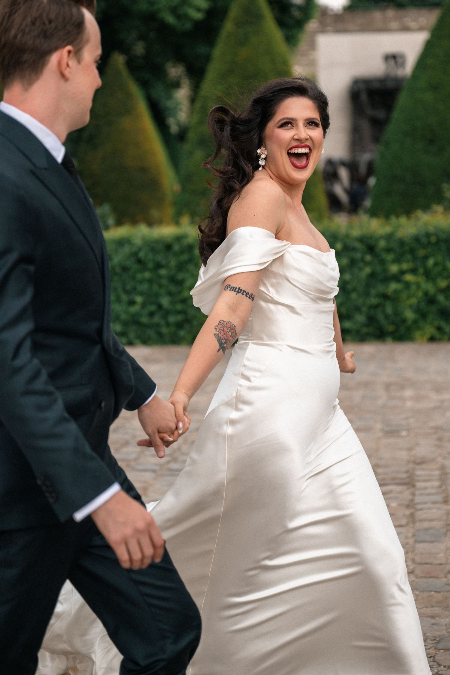 Joyful bride in off-shoulder white satin gown laughs while holding groom's hand walking through manicured garden.