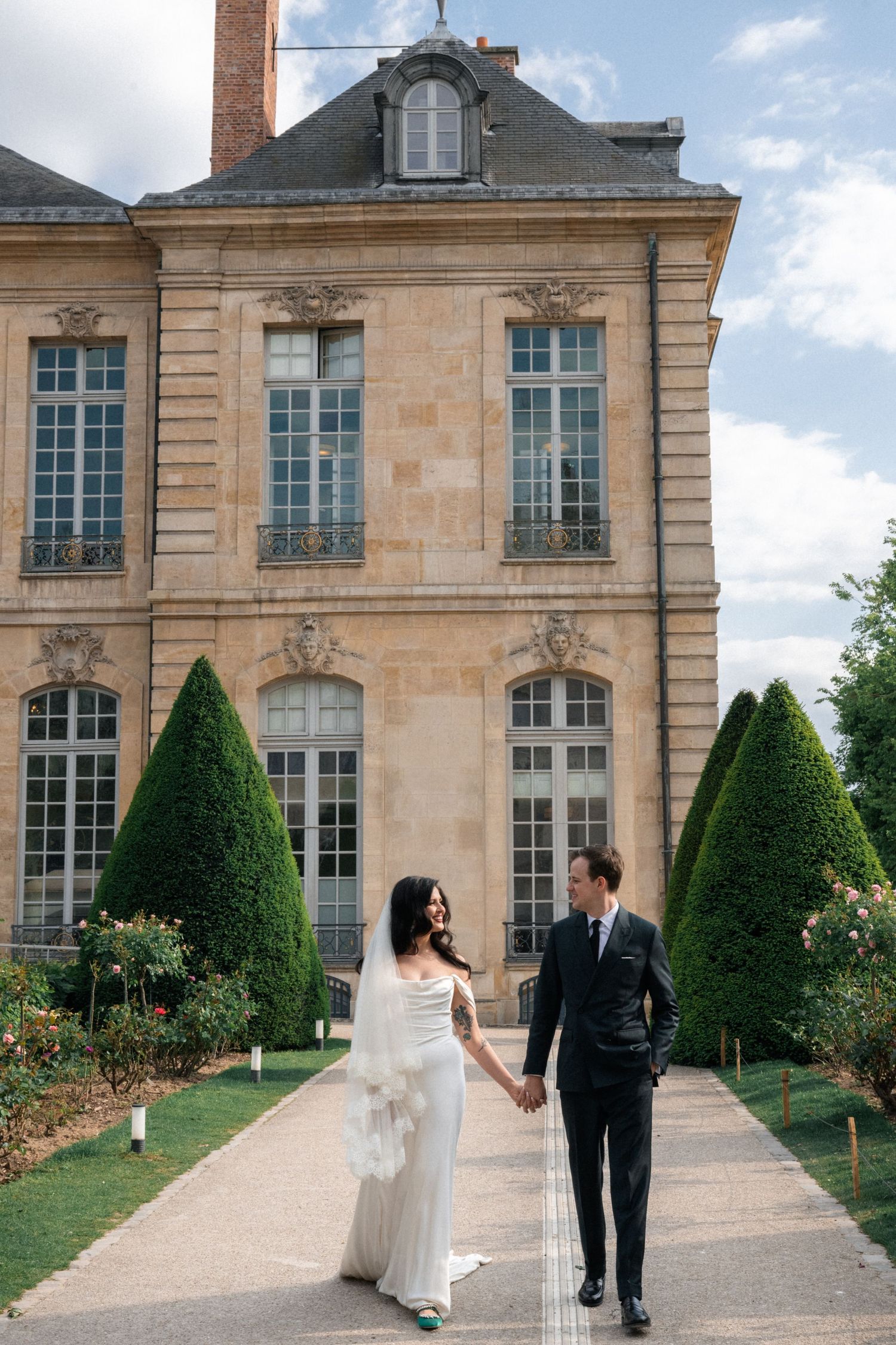 Wedding couple holding hands walking toward grand French chateau surrounded by manicured topiary gardens.