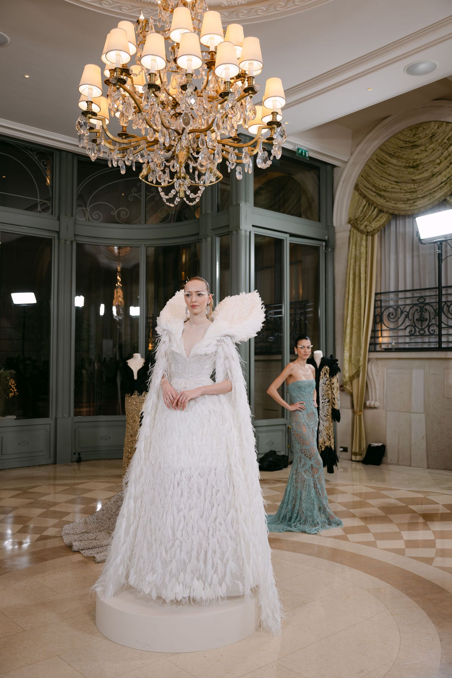 Model in elaborate white feathered wedding gown under a crystal chandelier in a luxurious ballroom.