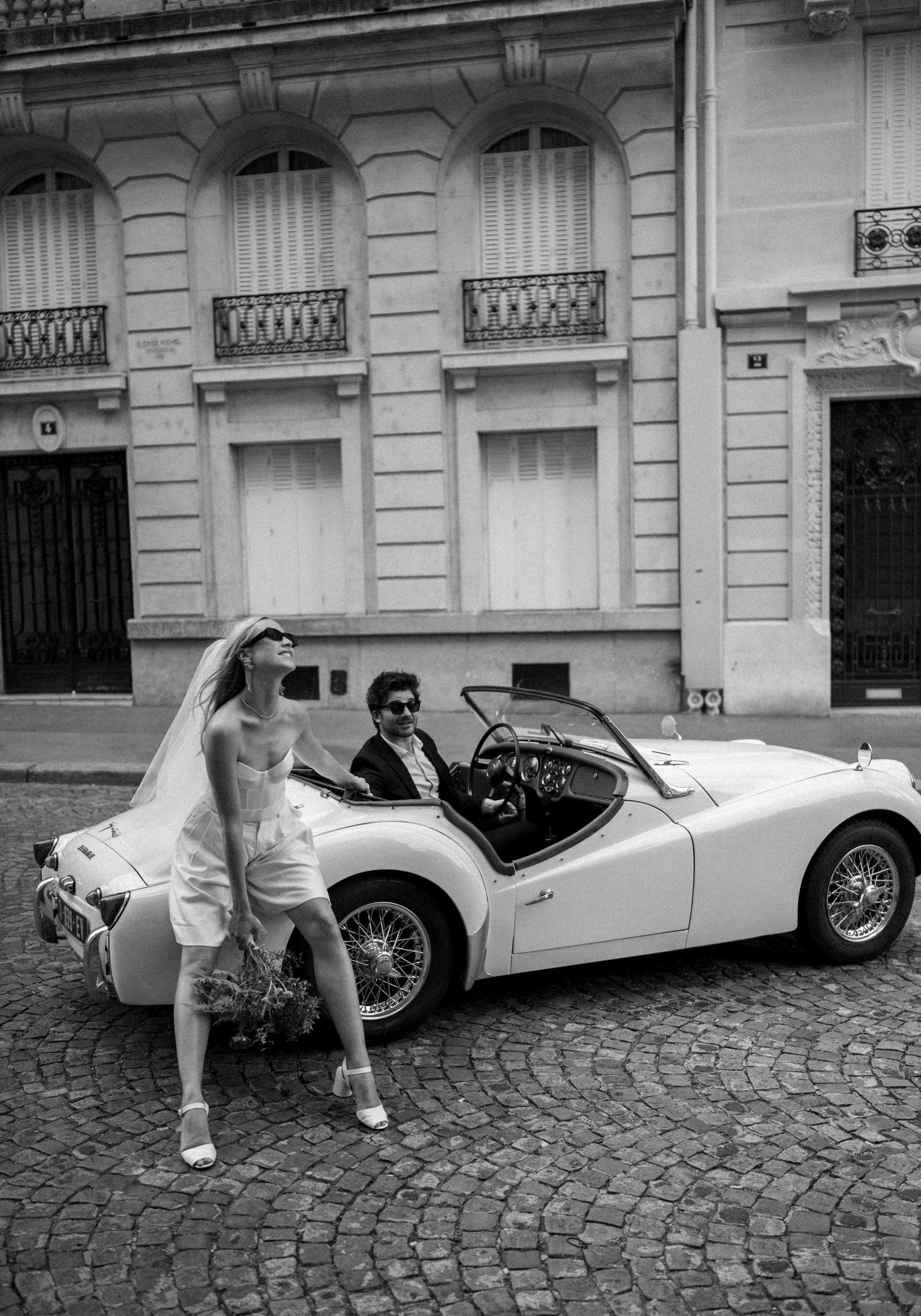 Bride in short white dress leans on vintage convertible as groom sits inside, black and white Paris wedding photo.