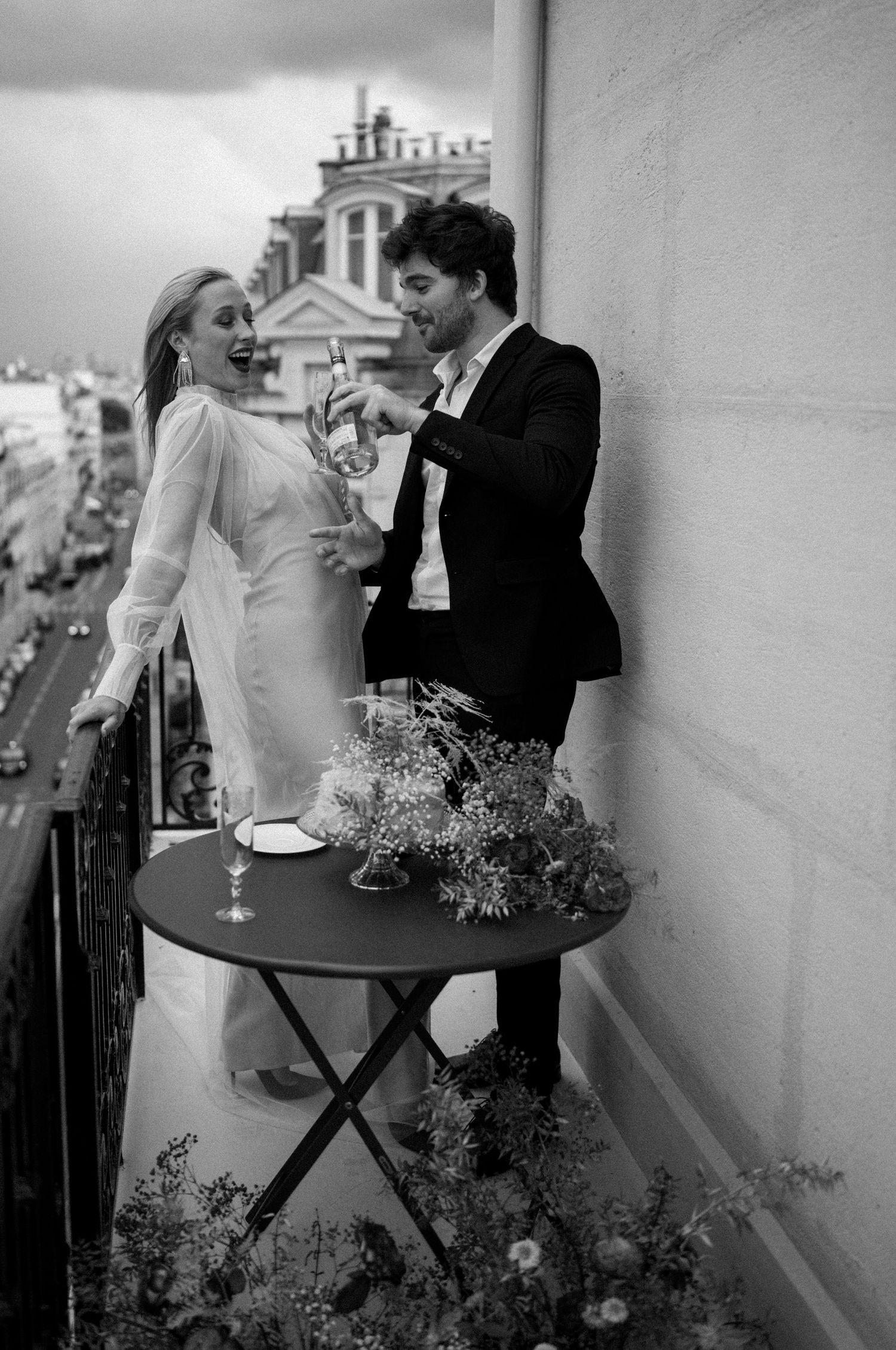 Black and white photo of laughing couple pouring champagne on a Parisian balcony with city views behind them.
