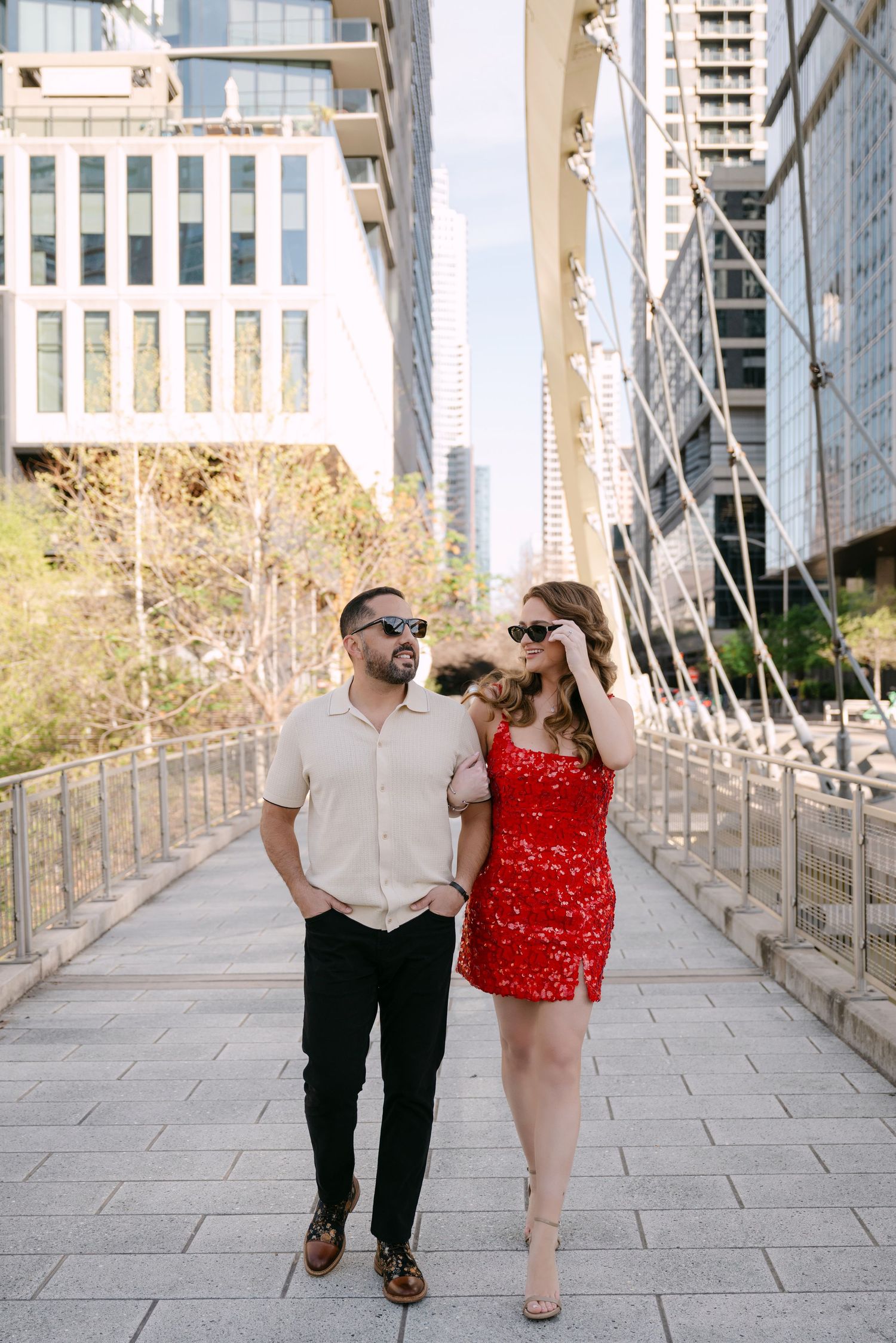 Couple in sunglasses walking between city skyscrapers, woman in red mini dress and man in beige shirt.