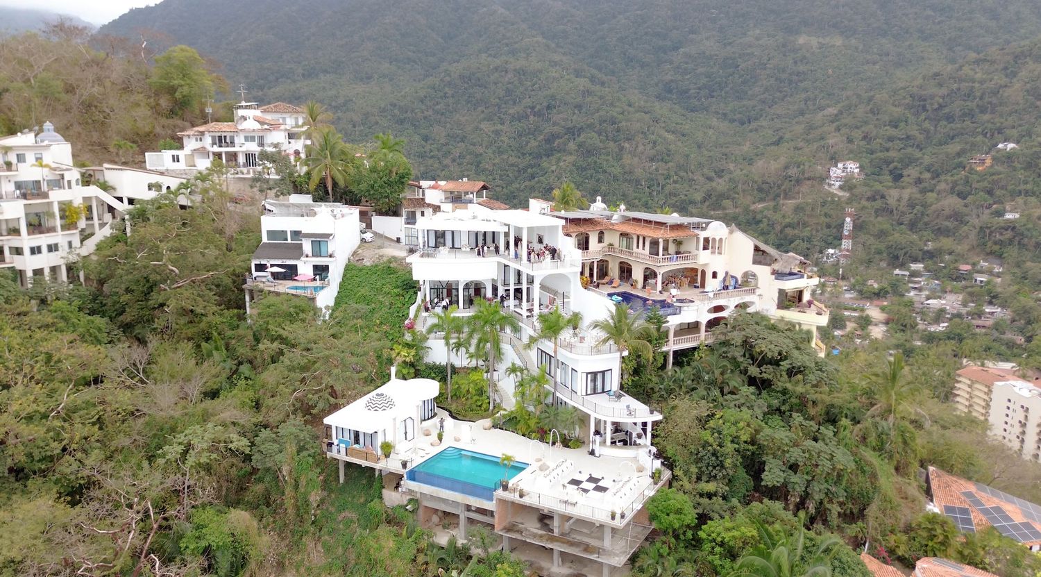 Aerial view of luxury white villas nestled in lush green hillside with mountain backdrop and pool.
