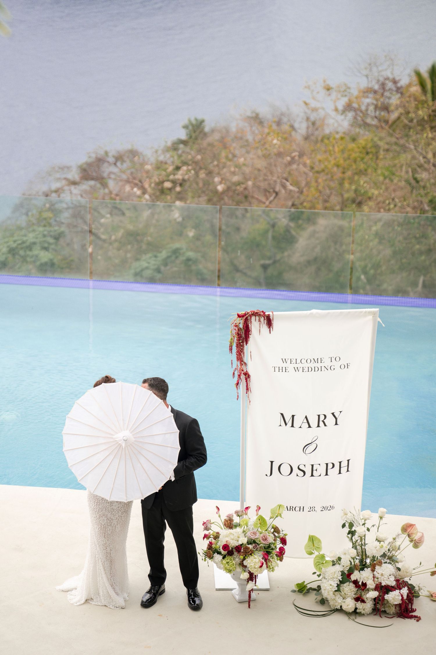 Bride and groom stand by a wedding sign reading Mary and Joseph beside floral arrangements near an infinity pool.