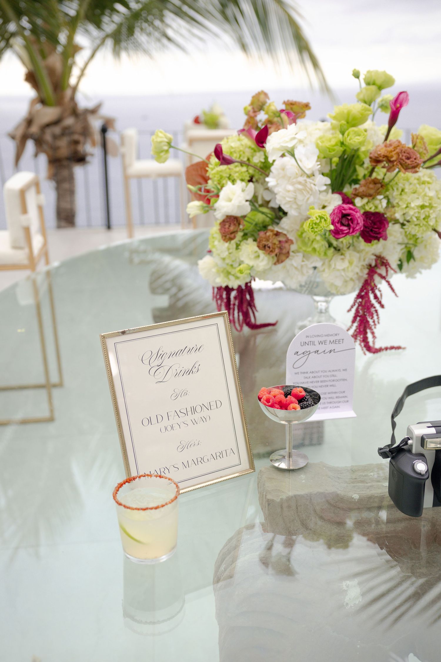 Elegant wedding signature drinks display with floral arrangement, cocktails, and framed menu sign on glass table.