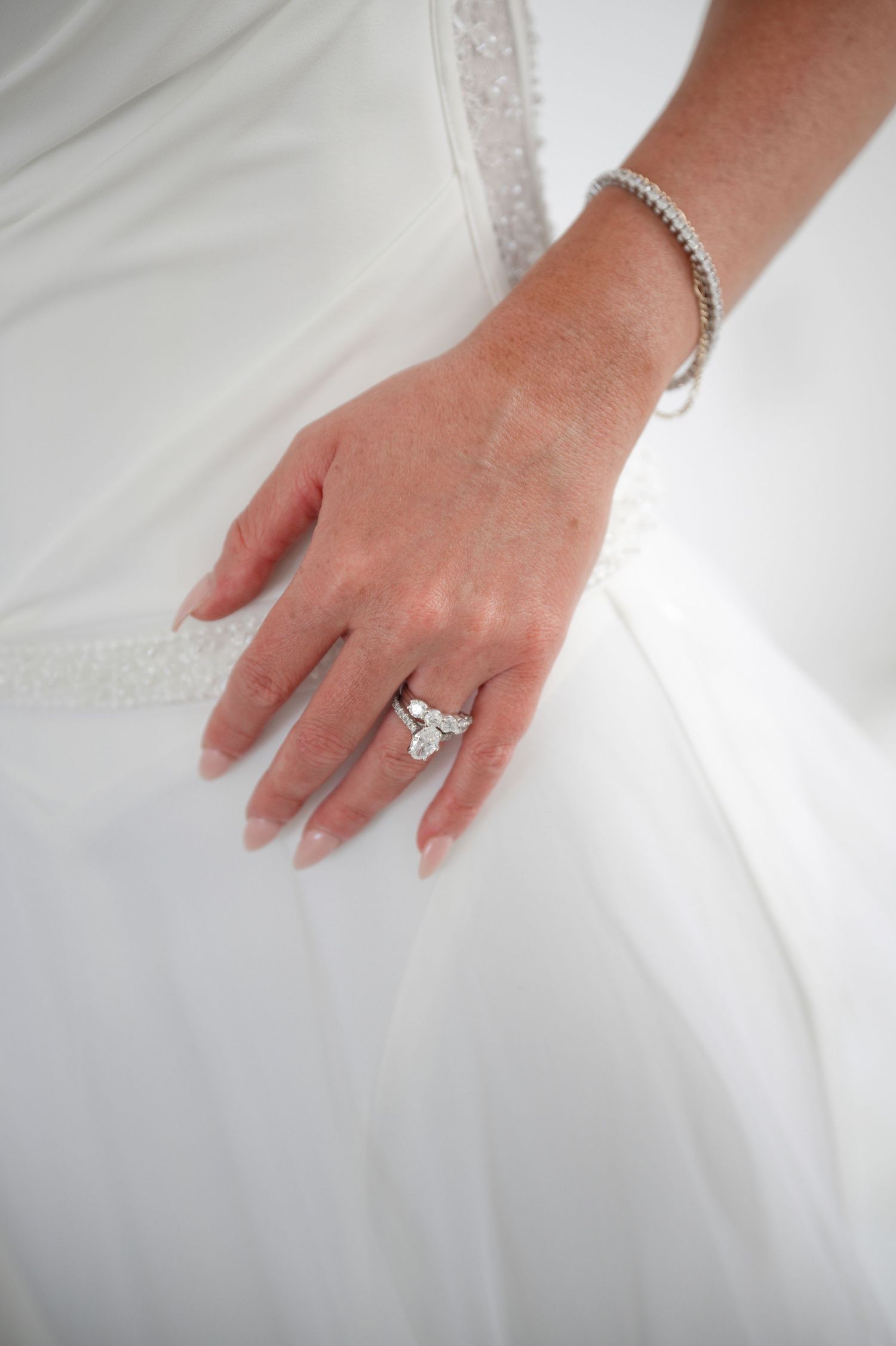 Close-up of bride's hand resting on white wedding gown showcasing a stunning diamond engagement ring and bracelet.