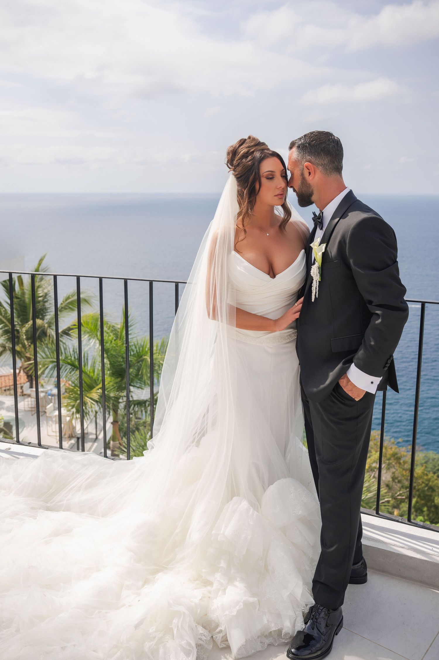 Wedding couple embraces on a tropical balcony overlooking lush greenery and blue ocean waters.