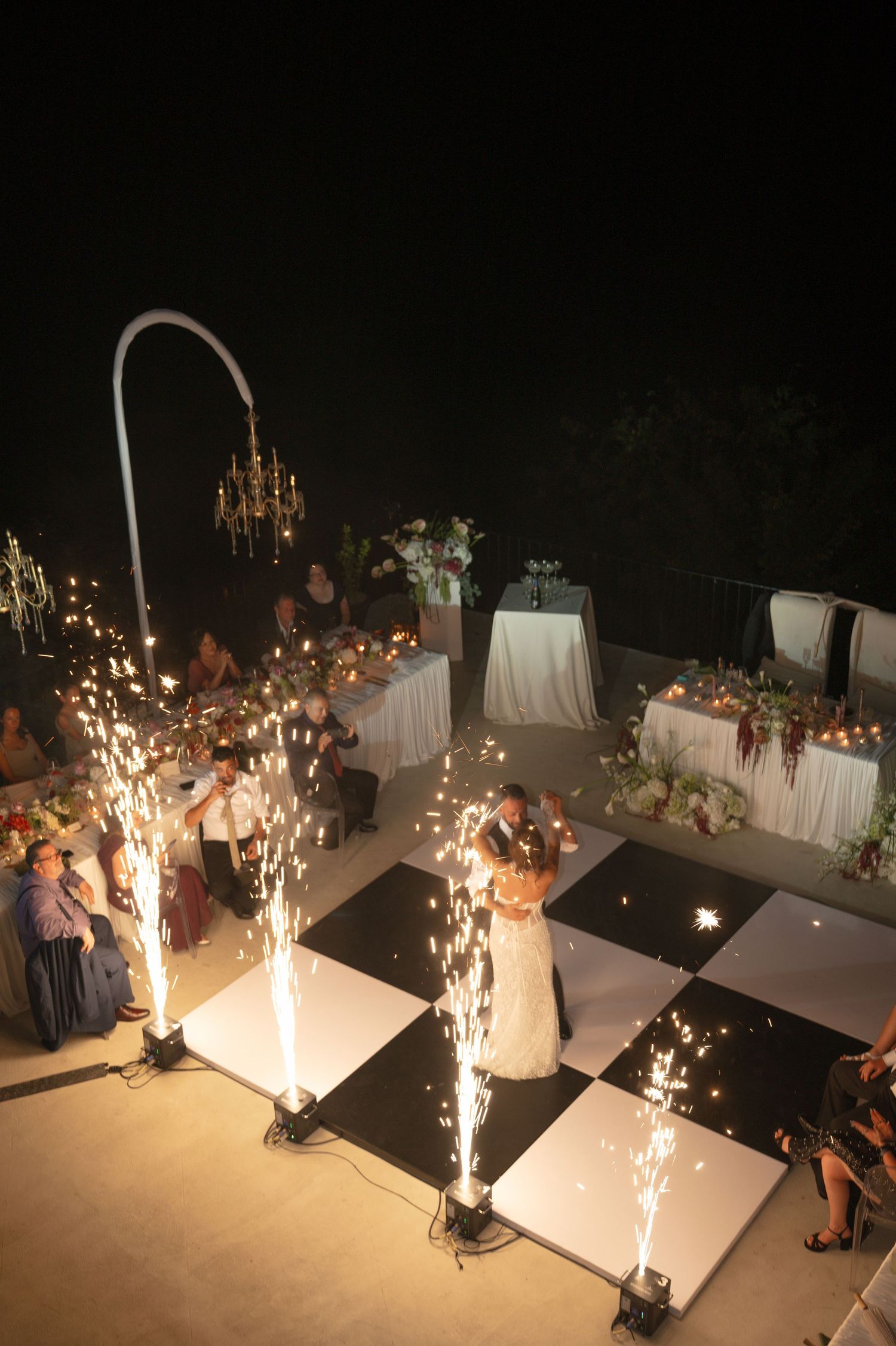 Bride and groom share first dance on checkered floor with sparklers at outdoor evening wedding reception.