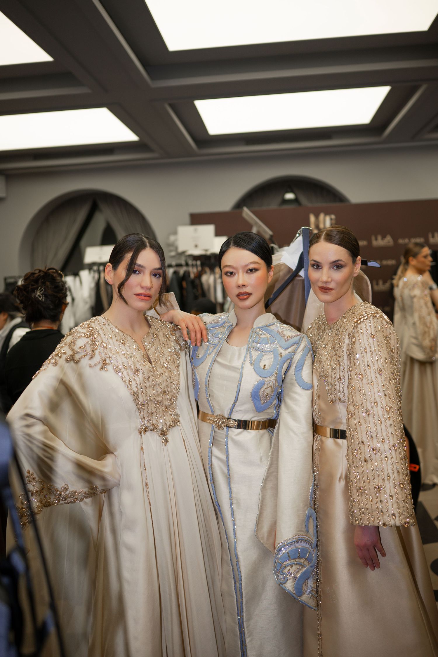Three models pose backstage in ornate embroidered gowns at a fashion show, wearing elegant cream and blue ensembles.
