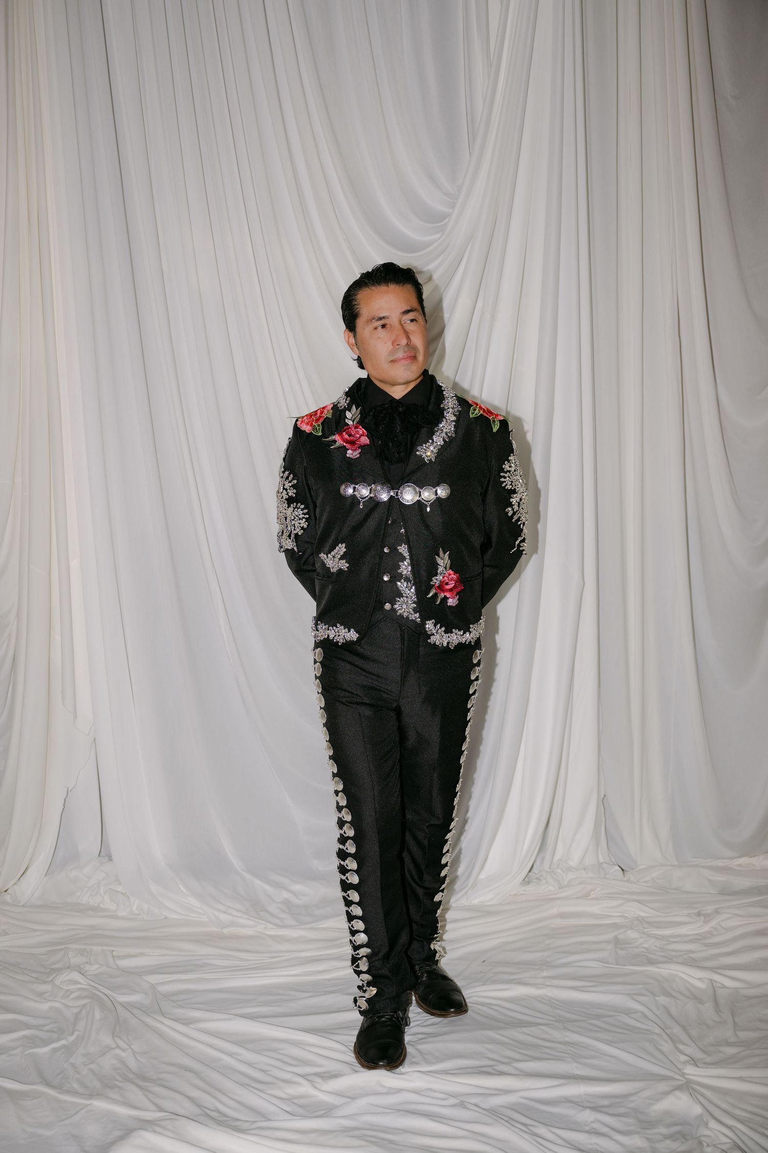 Man in black embroidered mariachi suit posing against white draped fabric backdrop.