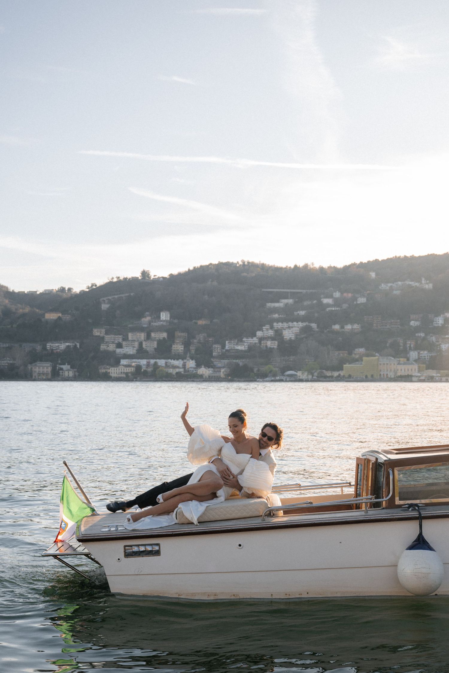 Bride and groom posing on a wooden boat on Lake Como, Italy, with scenic hillside village in the background.