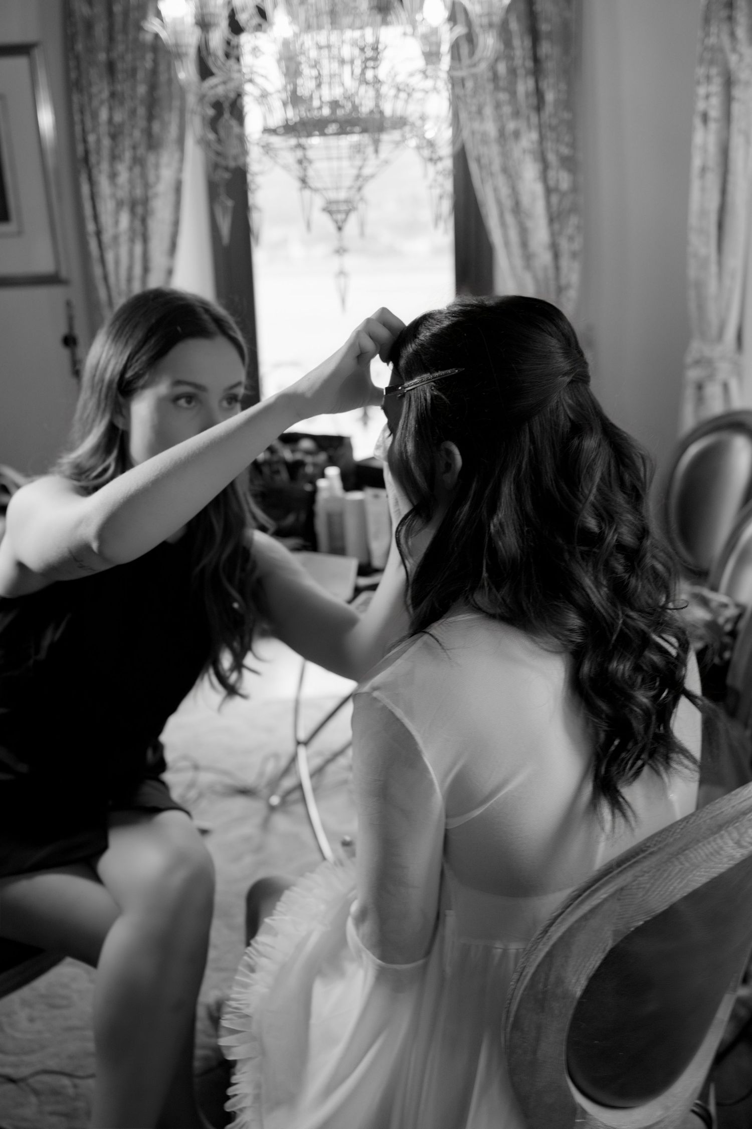 Black and white photo of a hairstylist styling a bride's dark curly hair in an elegant room with chandelier.