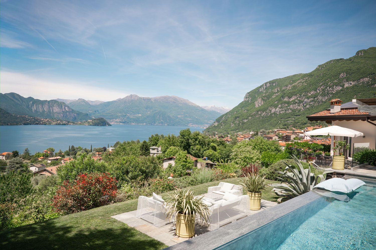 Luxury villa pool with panoramic views of Lake Como, lush gardens, and dramatic mountain backdrop on a sunny day.