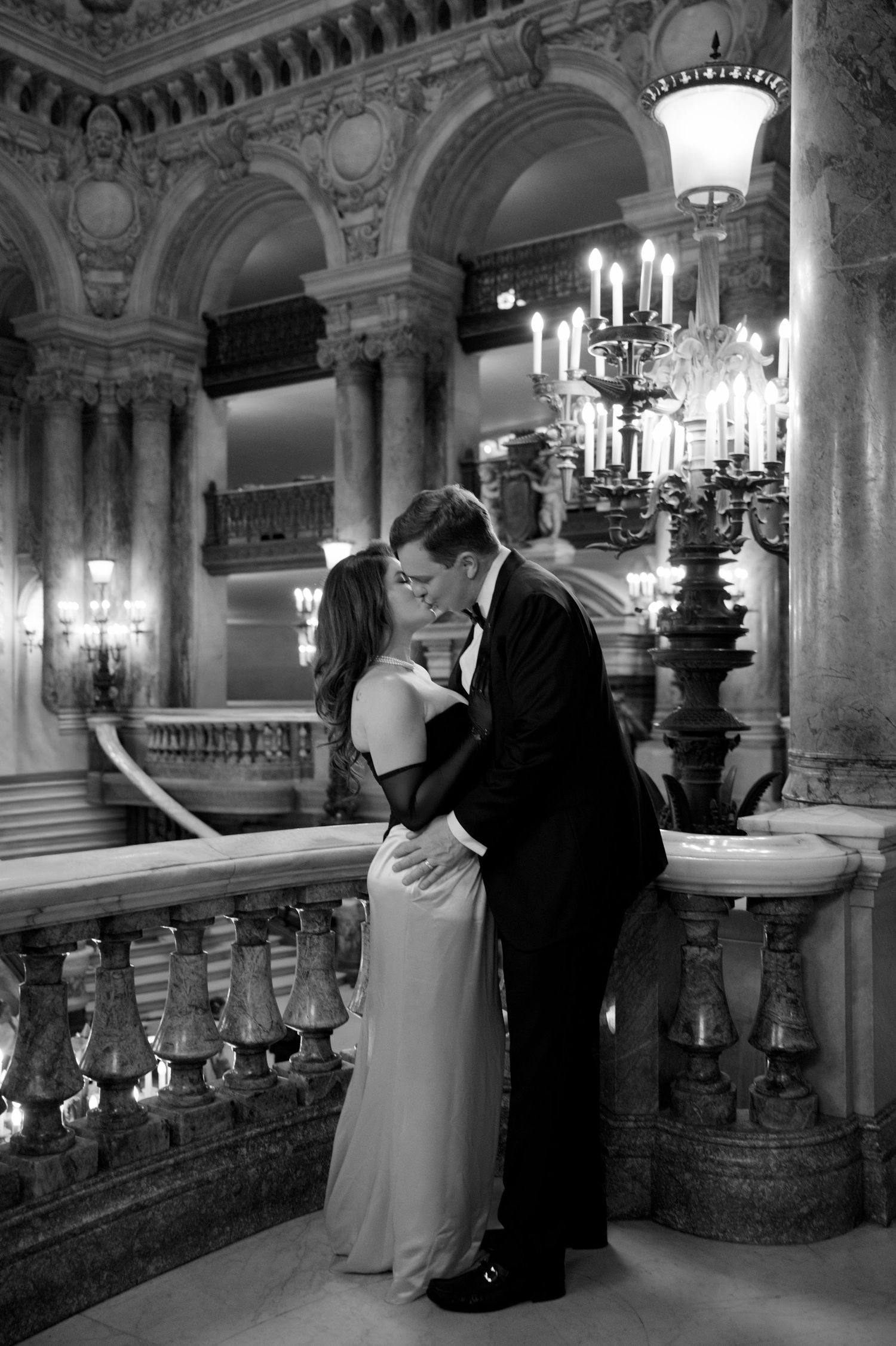 Elegant couple sharing a romantic kiss in a grand ballroom, black and white photo with ornate architecture and candelabras.