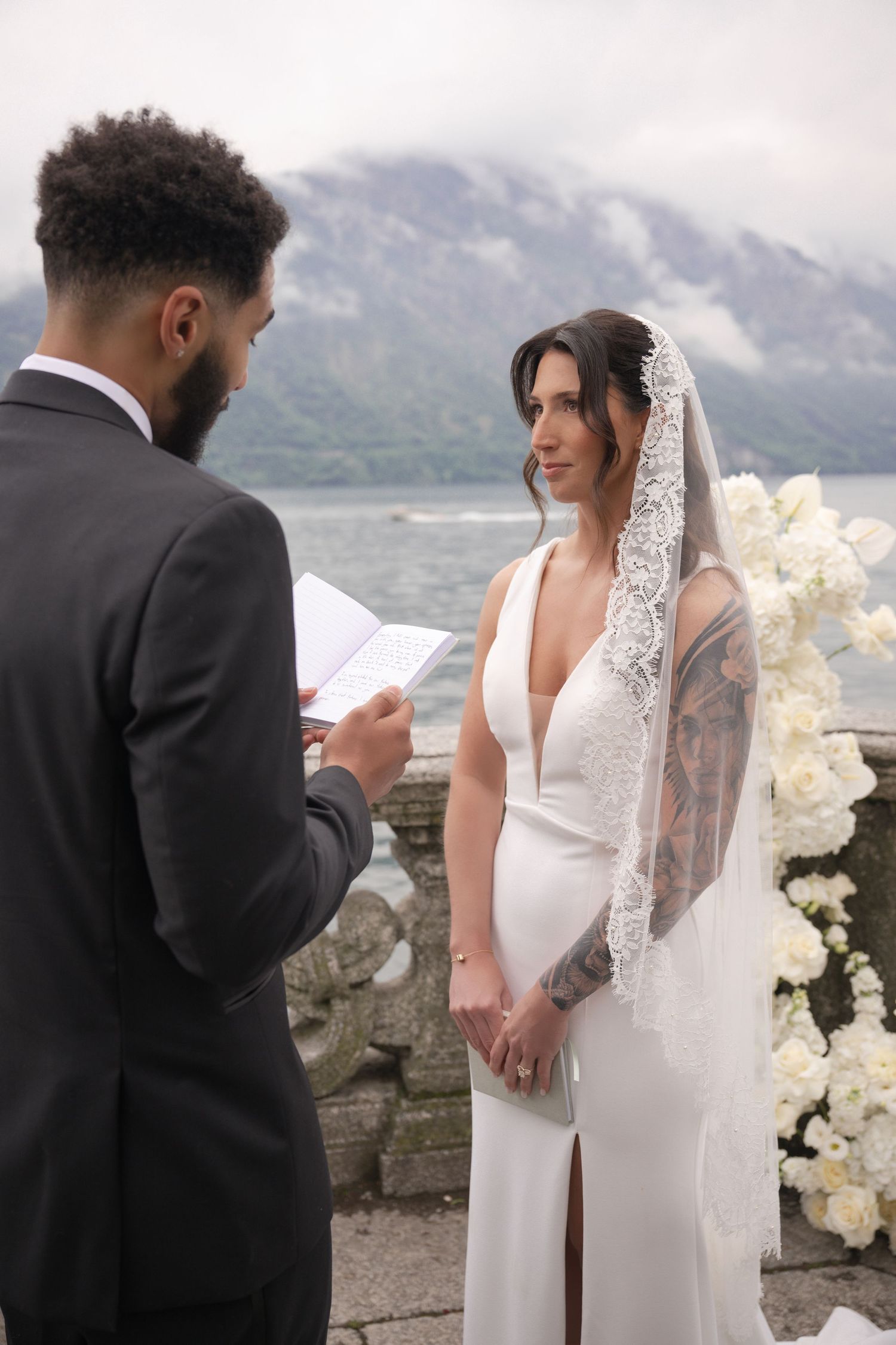 Groom reads wedding vows to bride in white gown and lace veil at lakeside ceremony with mountain backdrop.