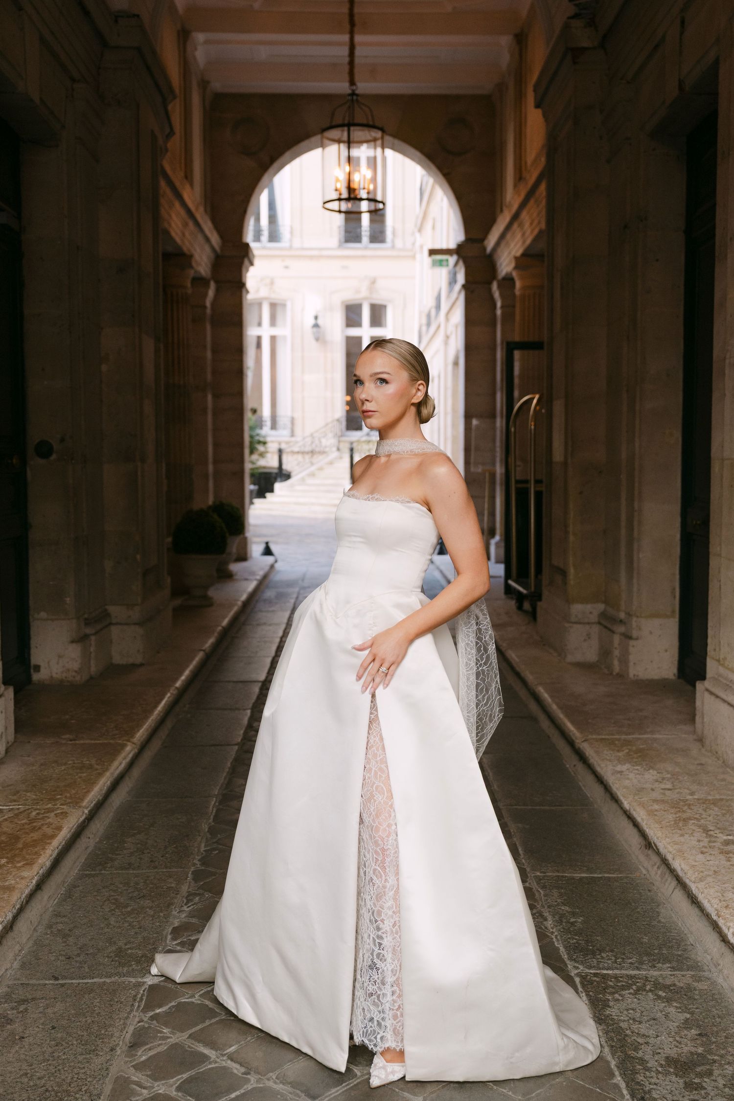 Elegant bride in strapless white gown with lace detail poses in a classic Parisian archway corridor.
