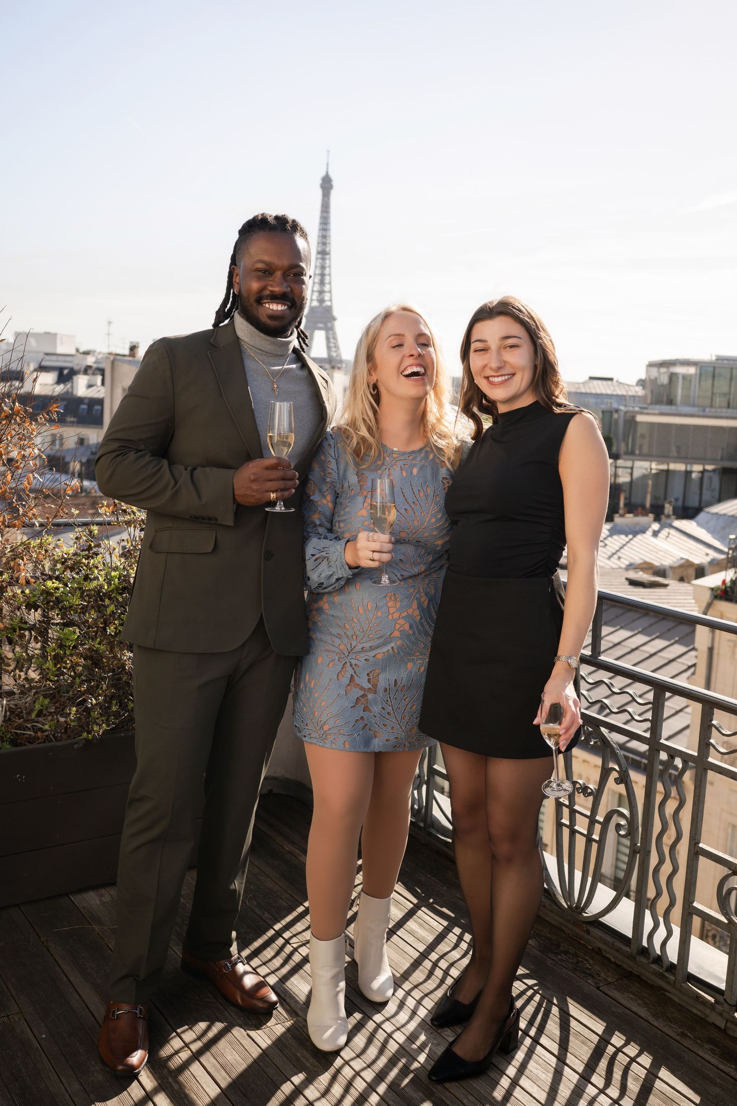 Three friends laugh and celebrate with champagne glasses on a Paris rooftop with the Eiffel Tower in the background.