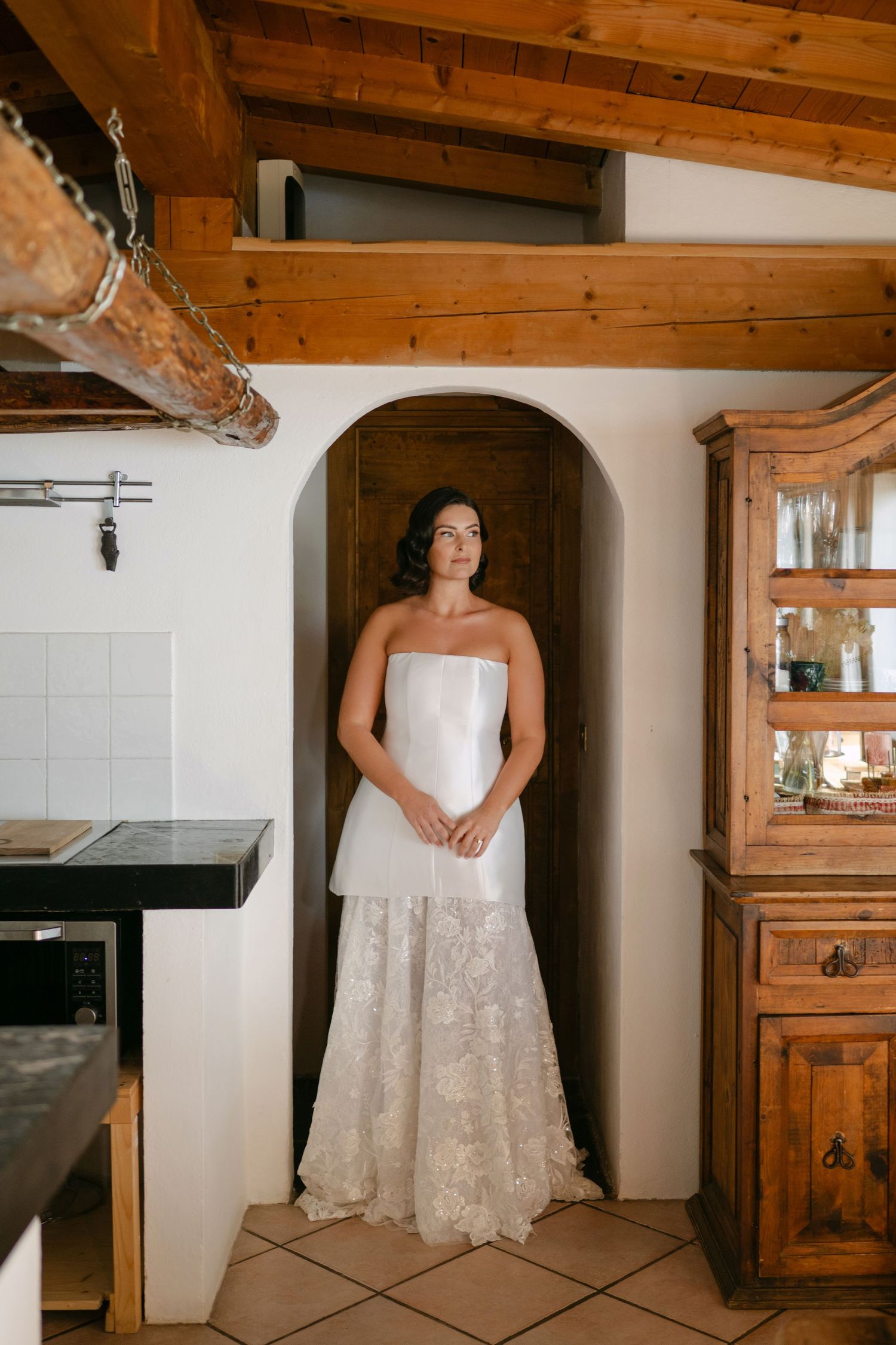 Bride in strapless white lace wedding gown standing in arched doorway of rustic kitchen with wooden beams.