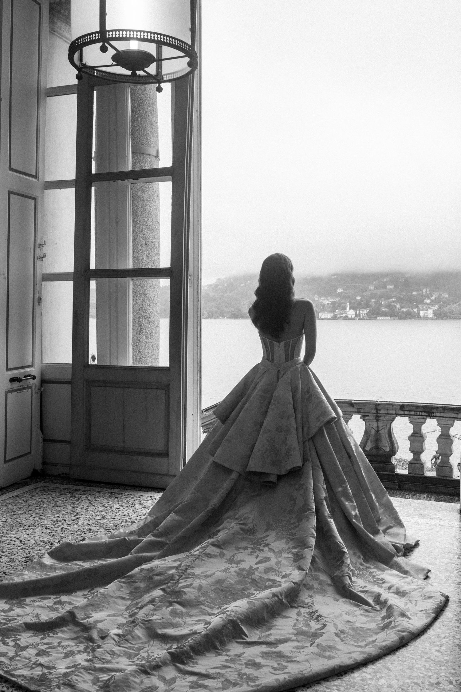 Black and white photo of a bride in an elegant ballgown with long train standing at an open door overlooking a lake.