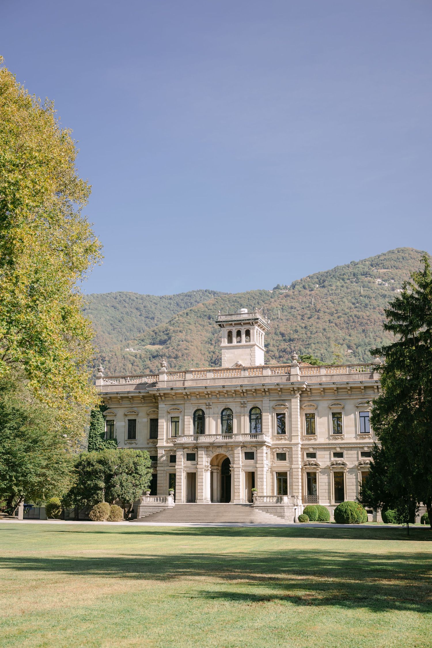 Elegant Italian villa with classical architecture set against green mountains and blue sky, surrounded by manicured lawns.