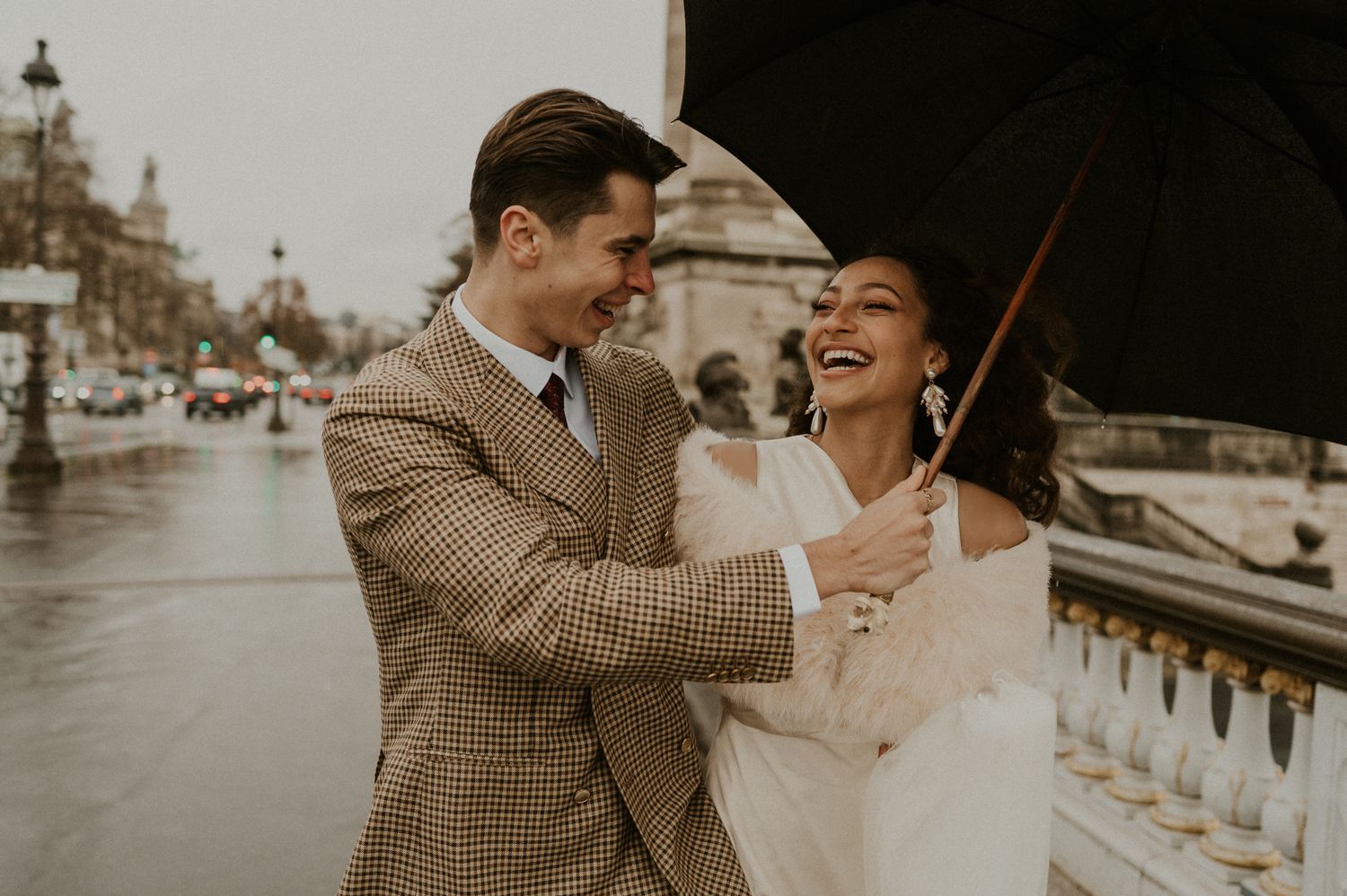 Joyful couple laughing together on a rainy day in Paris with ornate street lamps in the background.