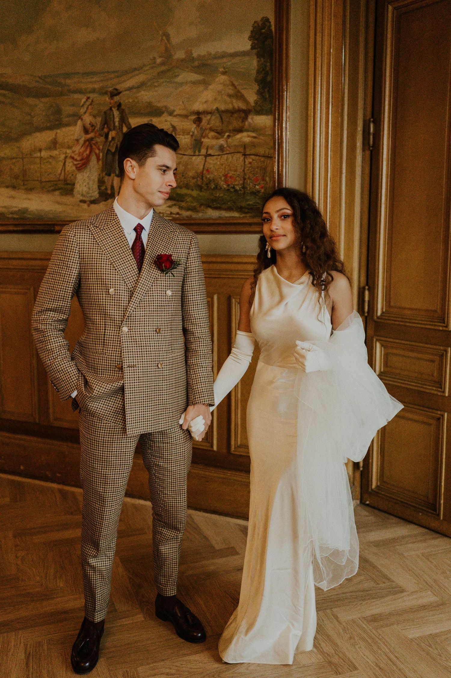 A couple in elegant wedding attire poses before a decorative wall mural in warm lighting.