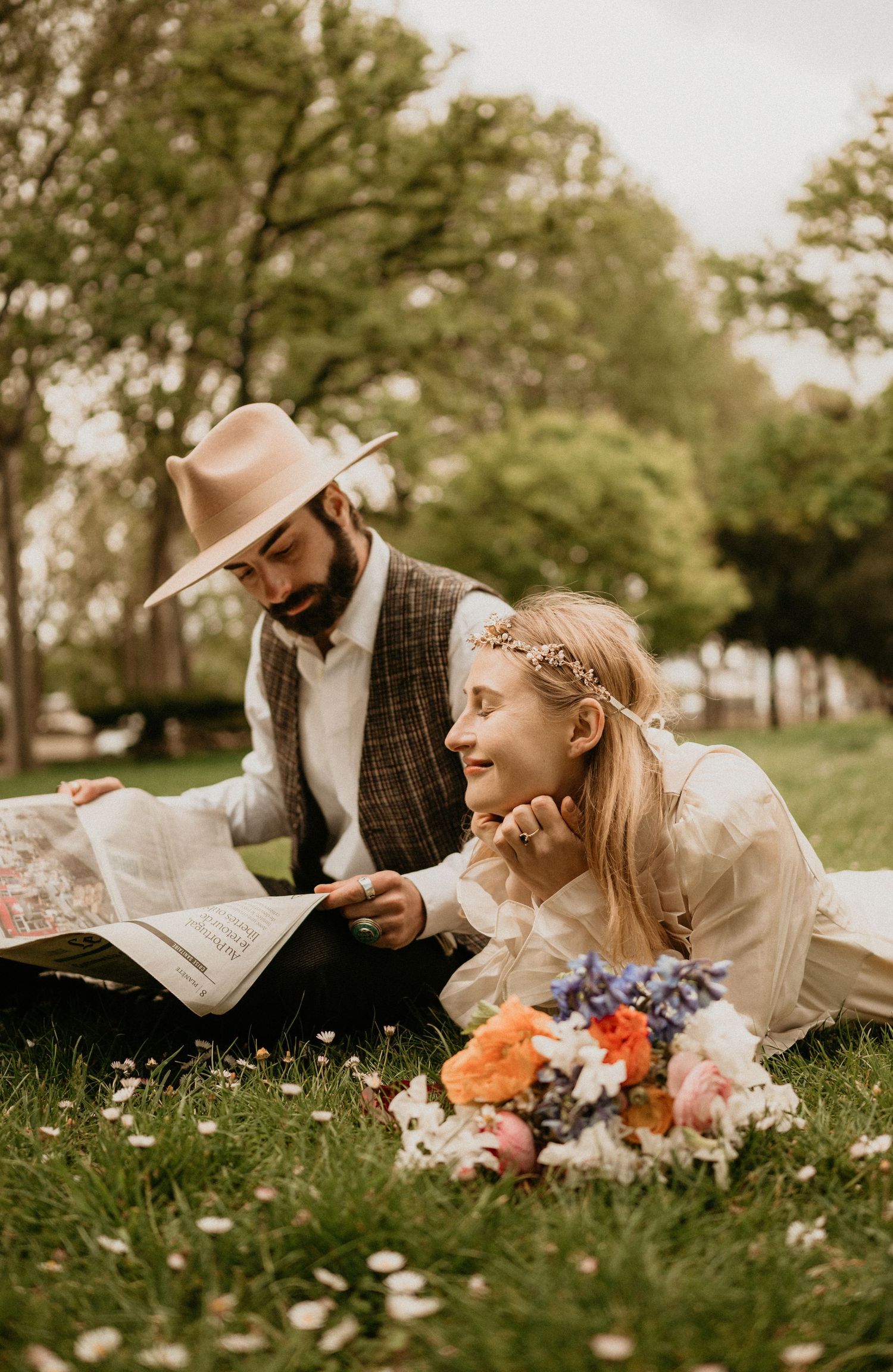 Couple sharing intimate moment while reading newspaper on grass with colorful bouquet nearby.