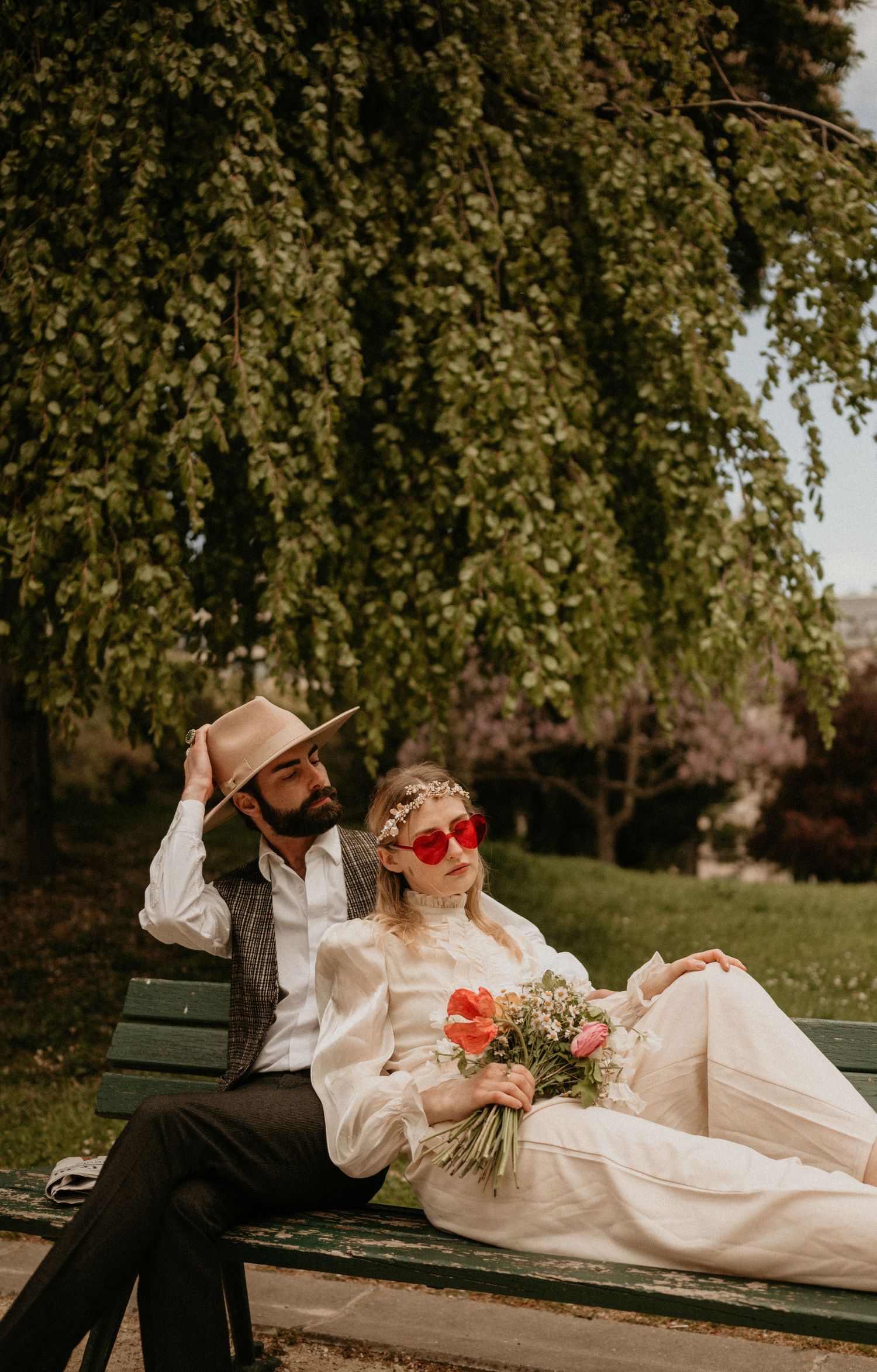 A romantic couple relaxes on a park bench under drooping willow branches in vintage-inspired attire with red heart sunglasses.