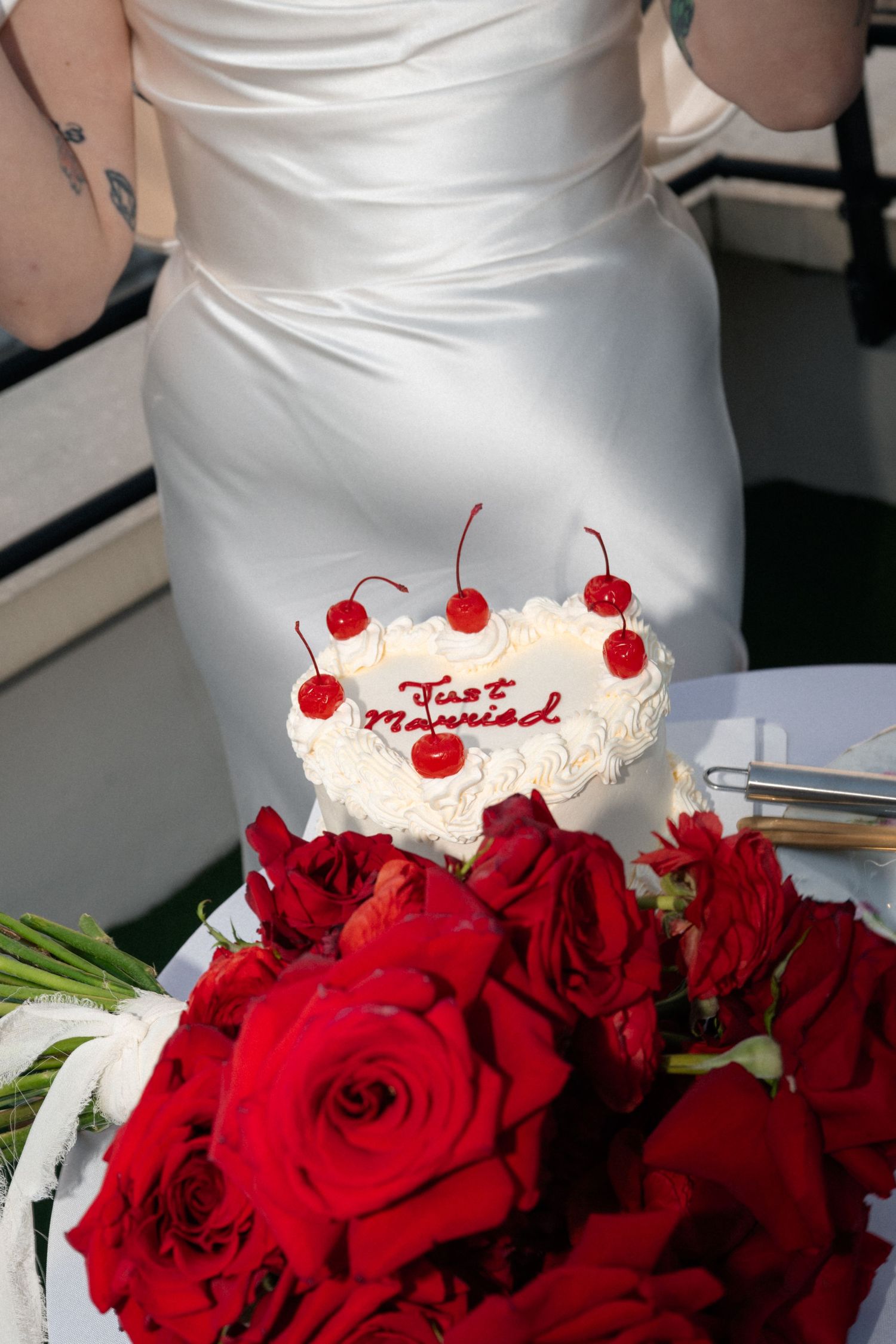 A white heart-shaped wedding cake decorated with cherries sits beside a bouquet of vibrant red roses.
