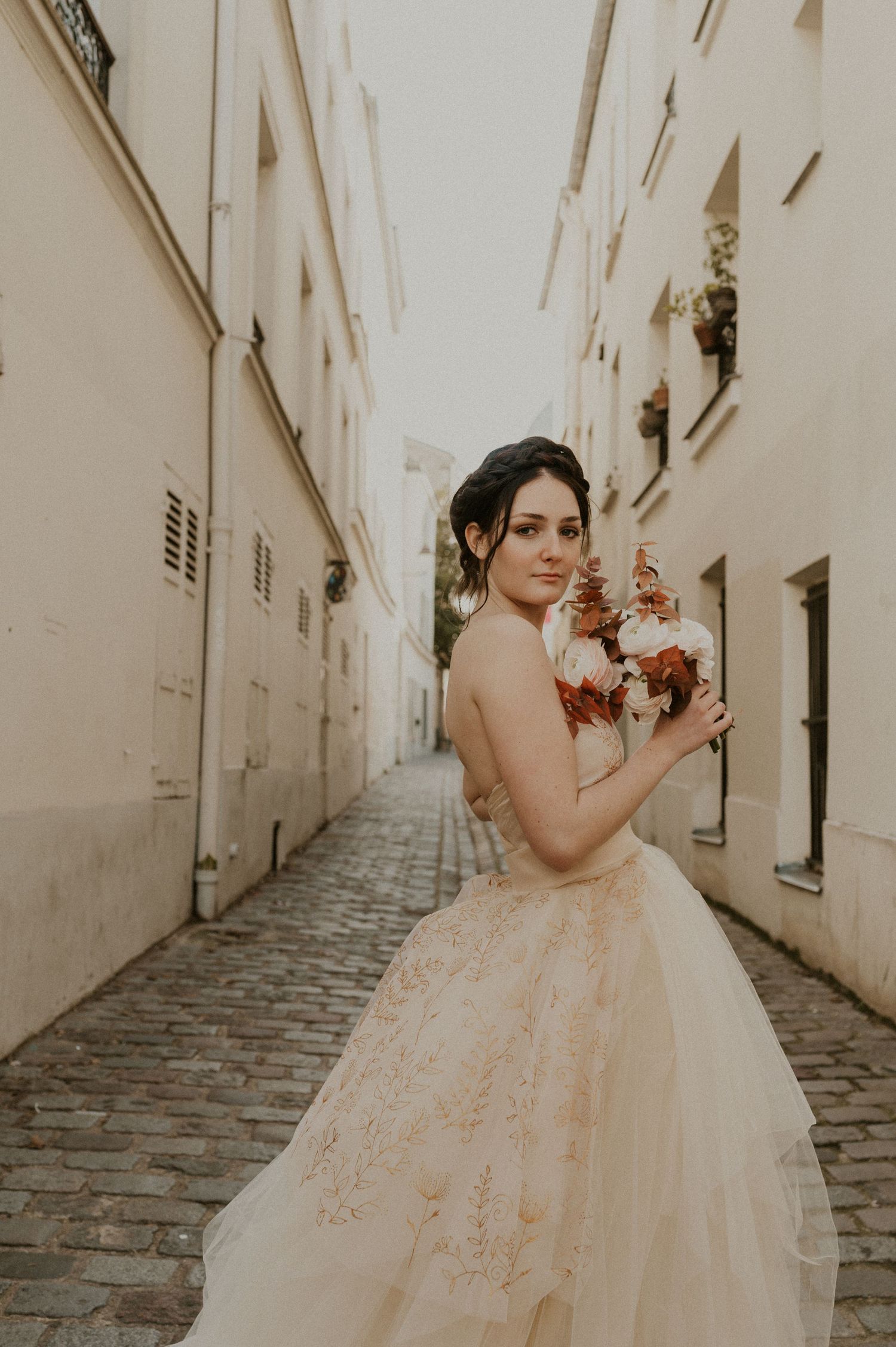 Bride holding autumn bouquet poses in strapless gown between white stone buildings.
