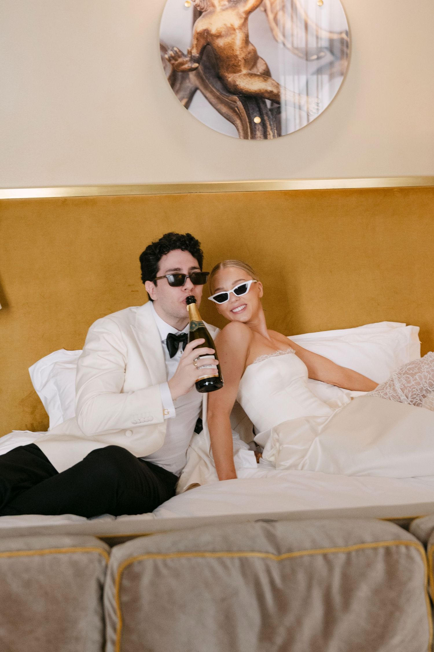 A couple in formal attire lies on white hotel bedding while looking at their phones.