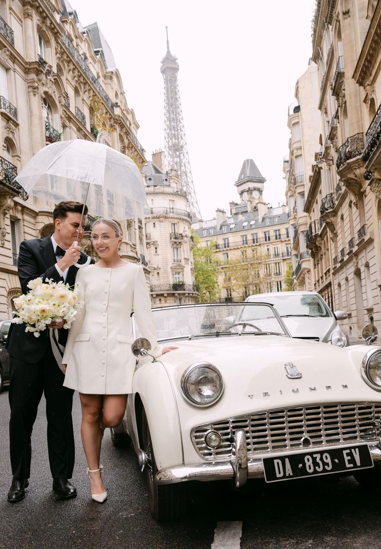 Vintage white Triumph car parked on cobblestone street in Paris with Eiffel Tower visible in background.