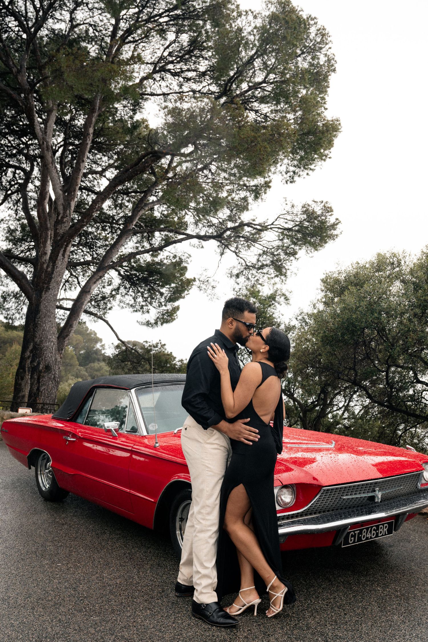 A couple sits together in a classic red car wearing black outfits and sunglasses on a sunny day.