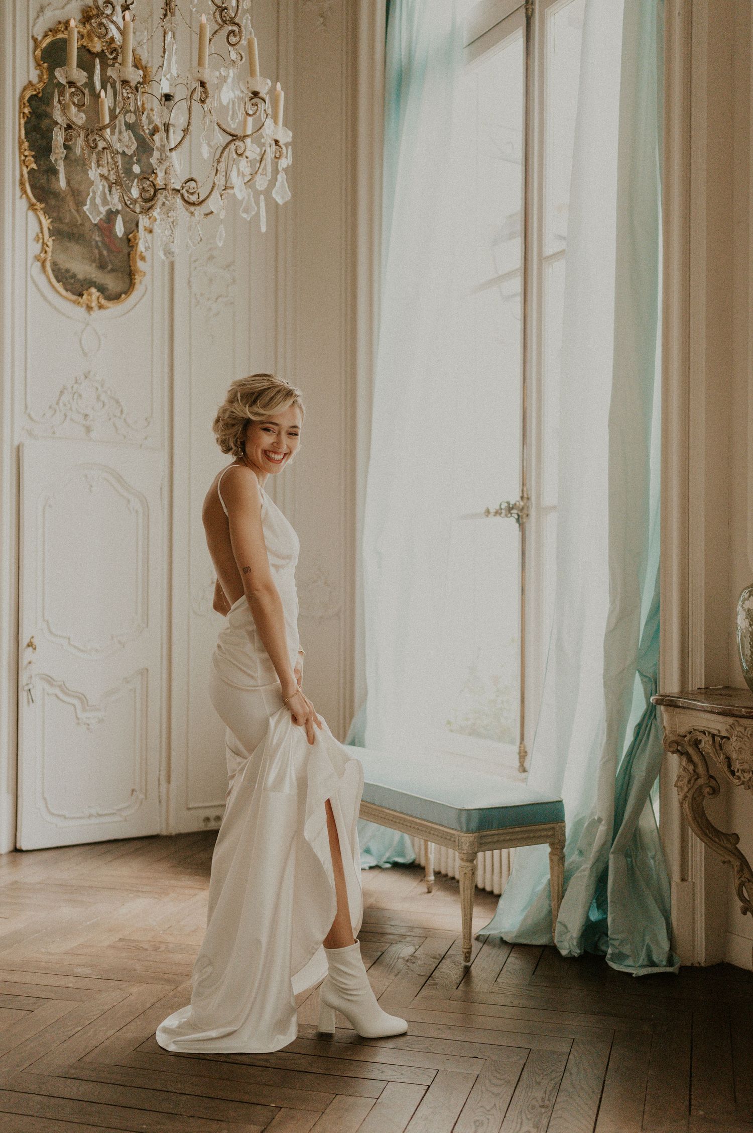 Elegant bride in backless white gown poses by window in ornate room with chandelier.