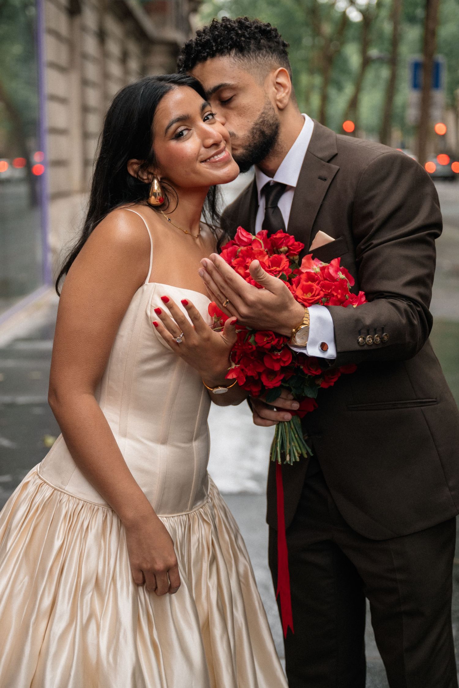Groom kissing bride holding red roses bouquet on a rainy Paris street.